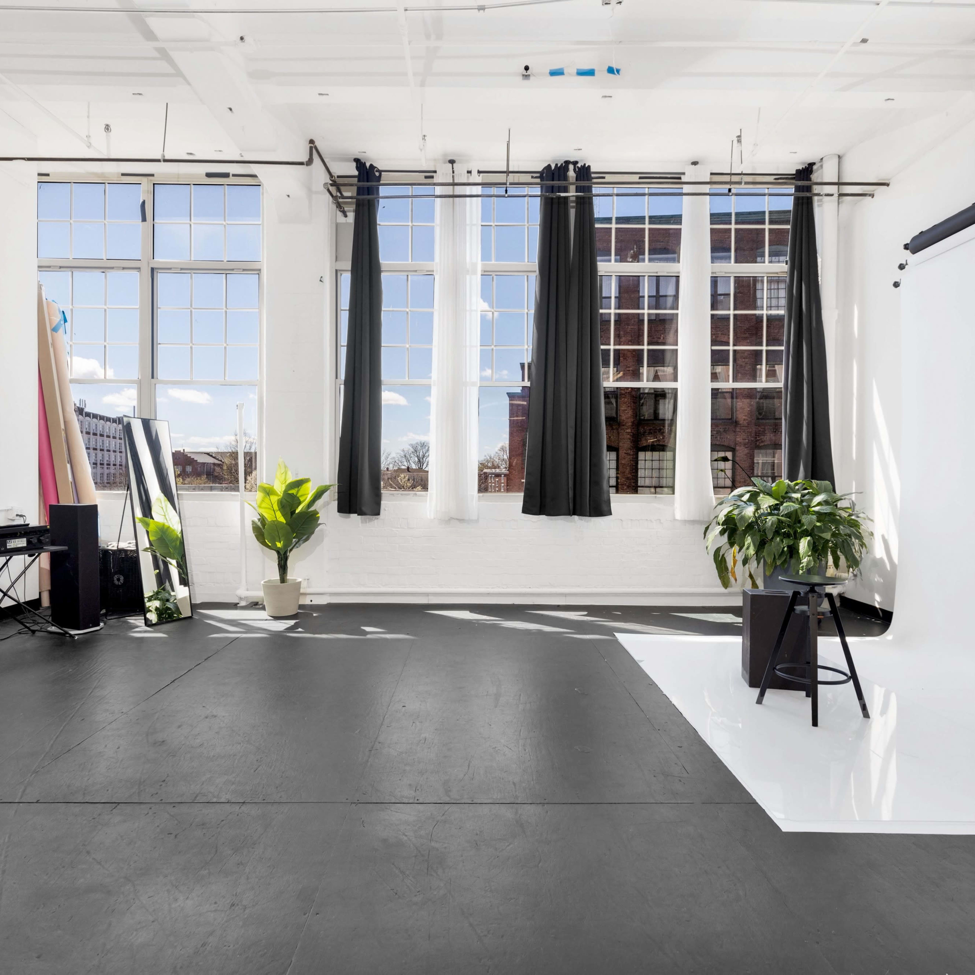 A bright, empty studio space with large windows, a black floor, and minimal furnishings, including a fan and potted plants.