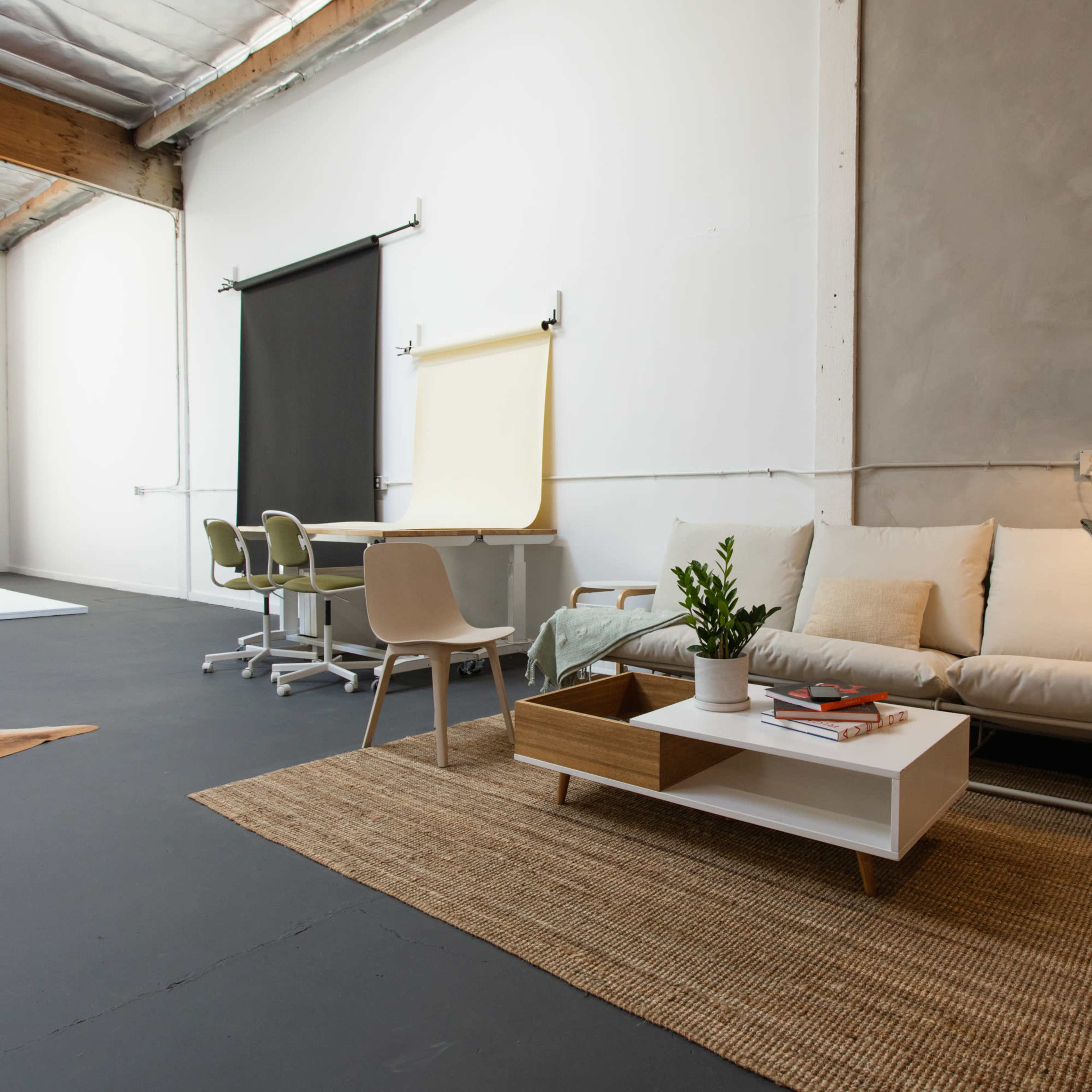 A modern open space with a seating area featuring a sofa, a coffee table, and a desk with chairs, all set on a dark floor with a large backdrop visible in the background.