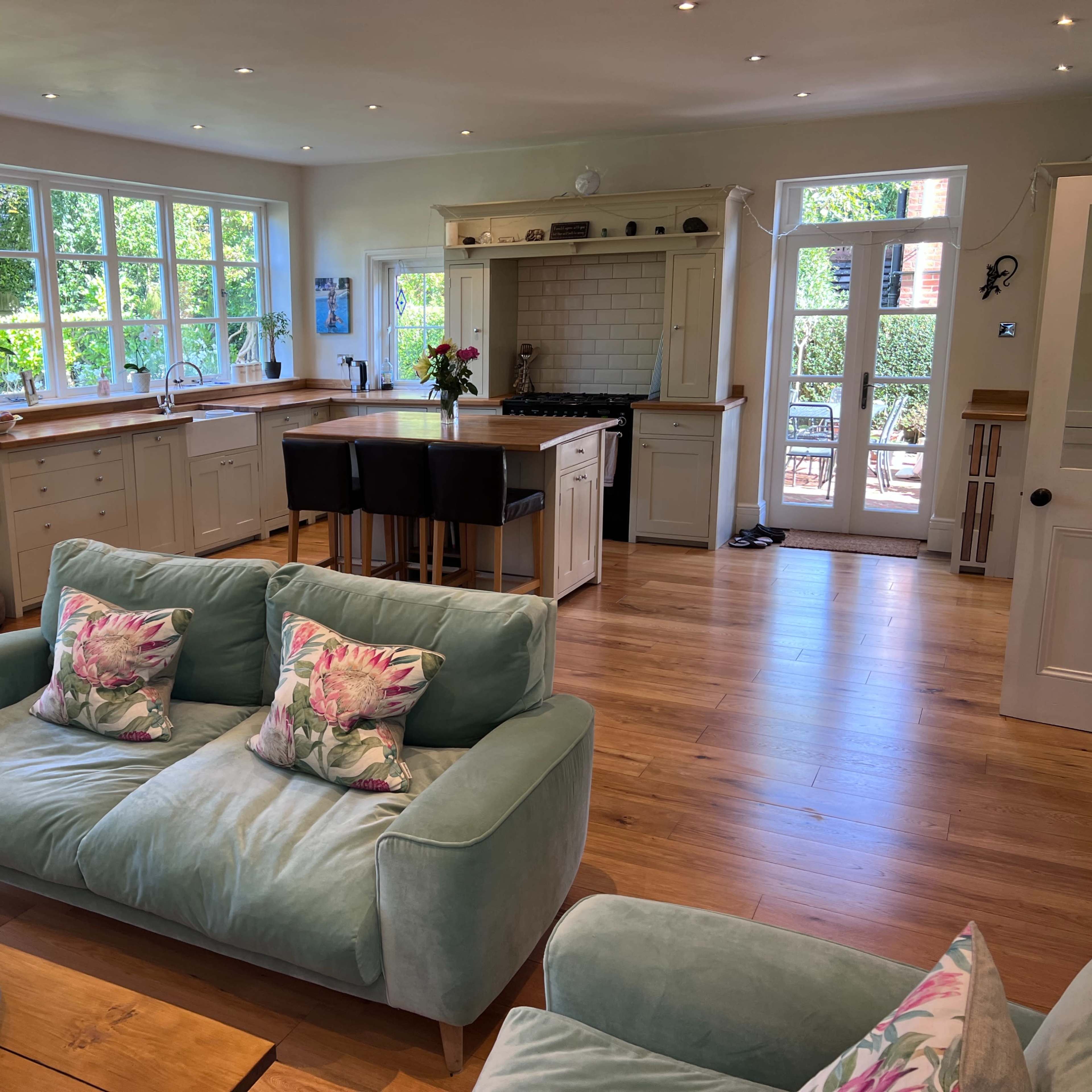 A bright, airy kitchen and living area with wooden floors, a large window, and a light-colored sofa with decorative cushions.