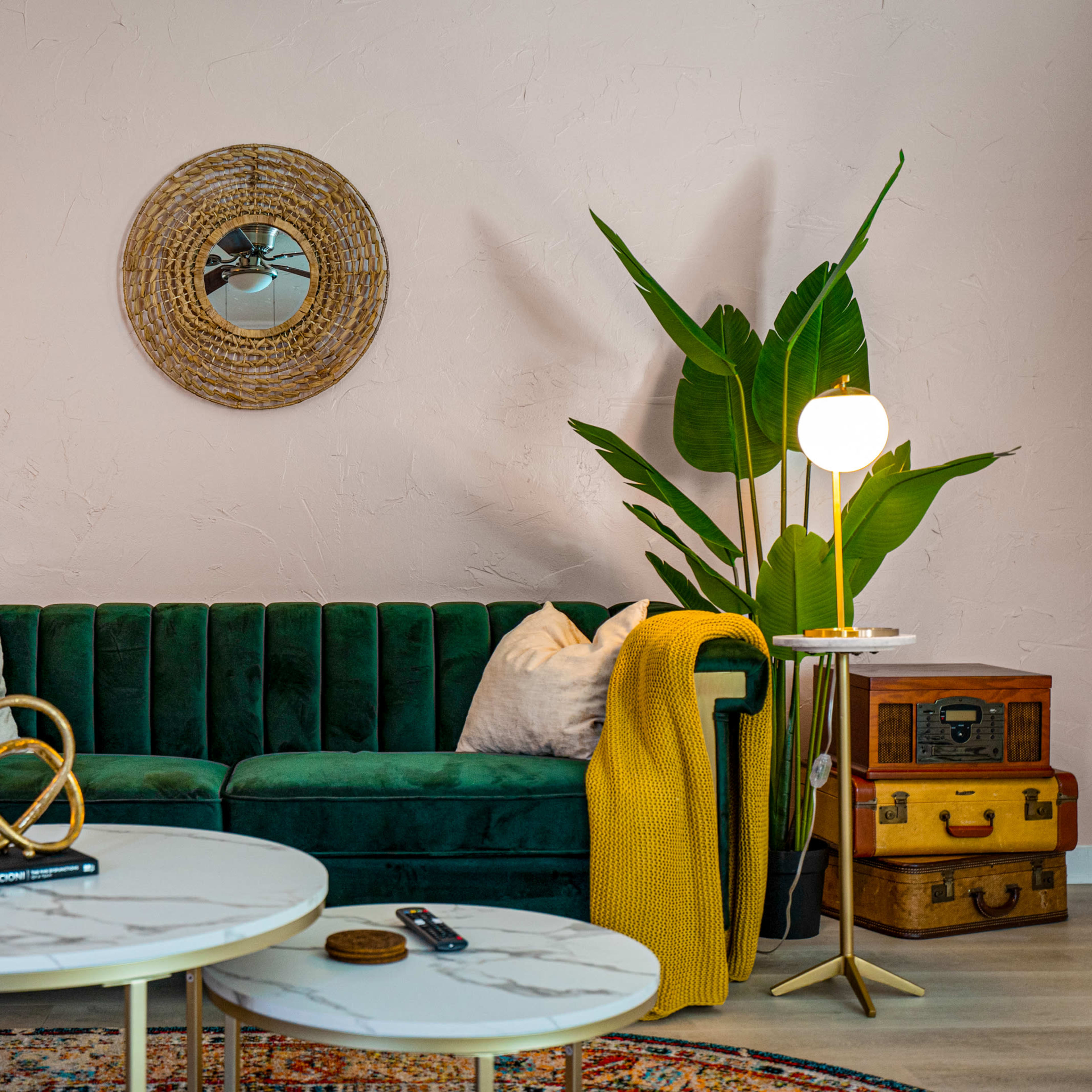 The scene shows a stylish living room featuring a green velvet sofa, a round mirror, potted plants, a marble coffee table, and accent decor on a light pink wall.