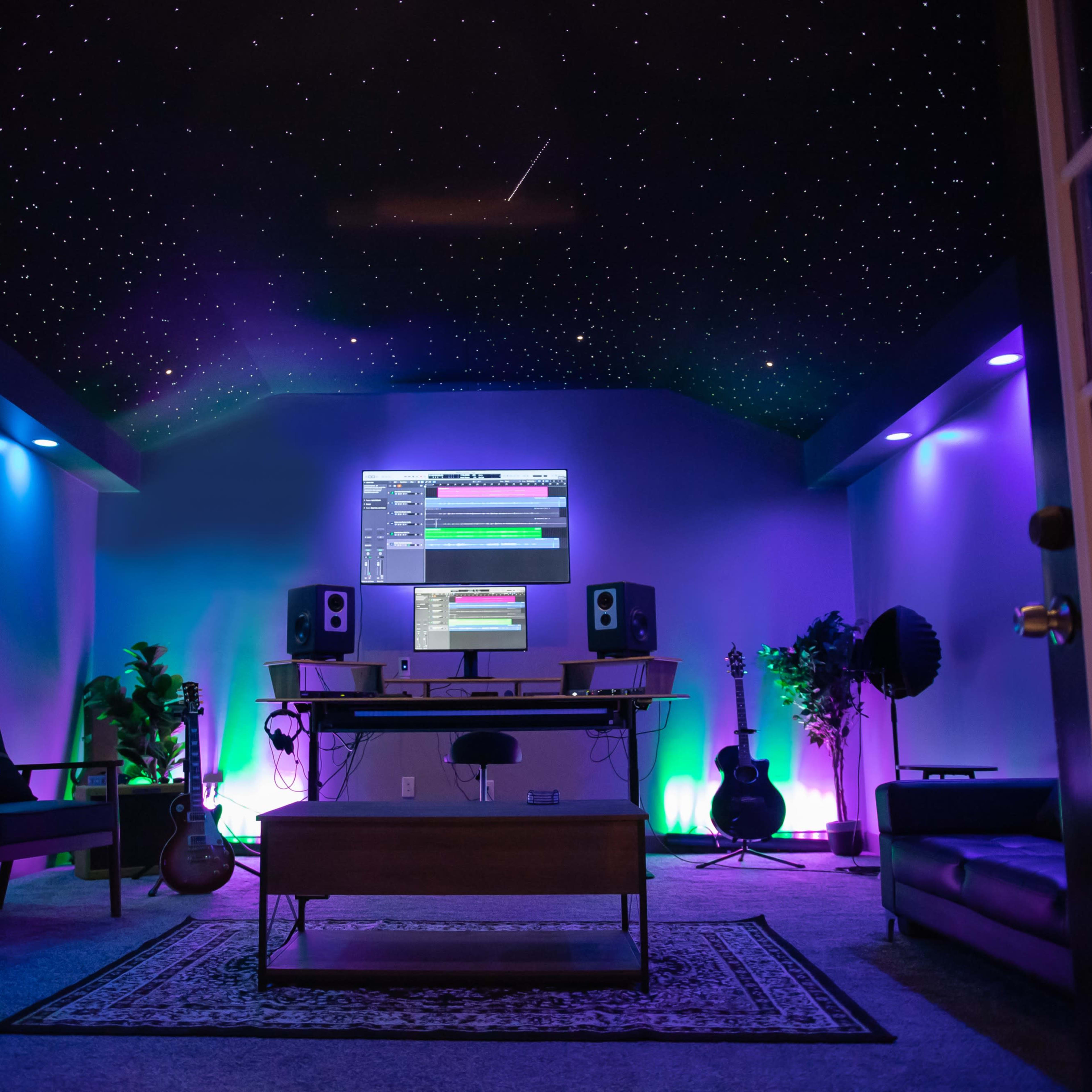 A music production room features a computer setup with studio monitors, illuminated plants, and colorful lighting under a ceiling resembling a starry sky.