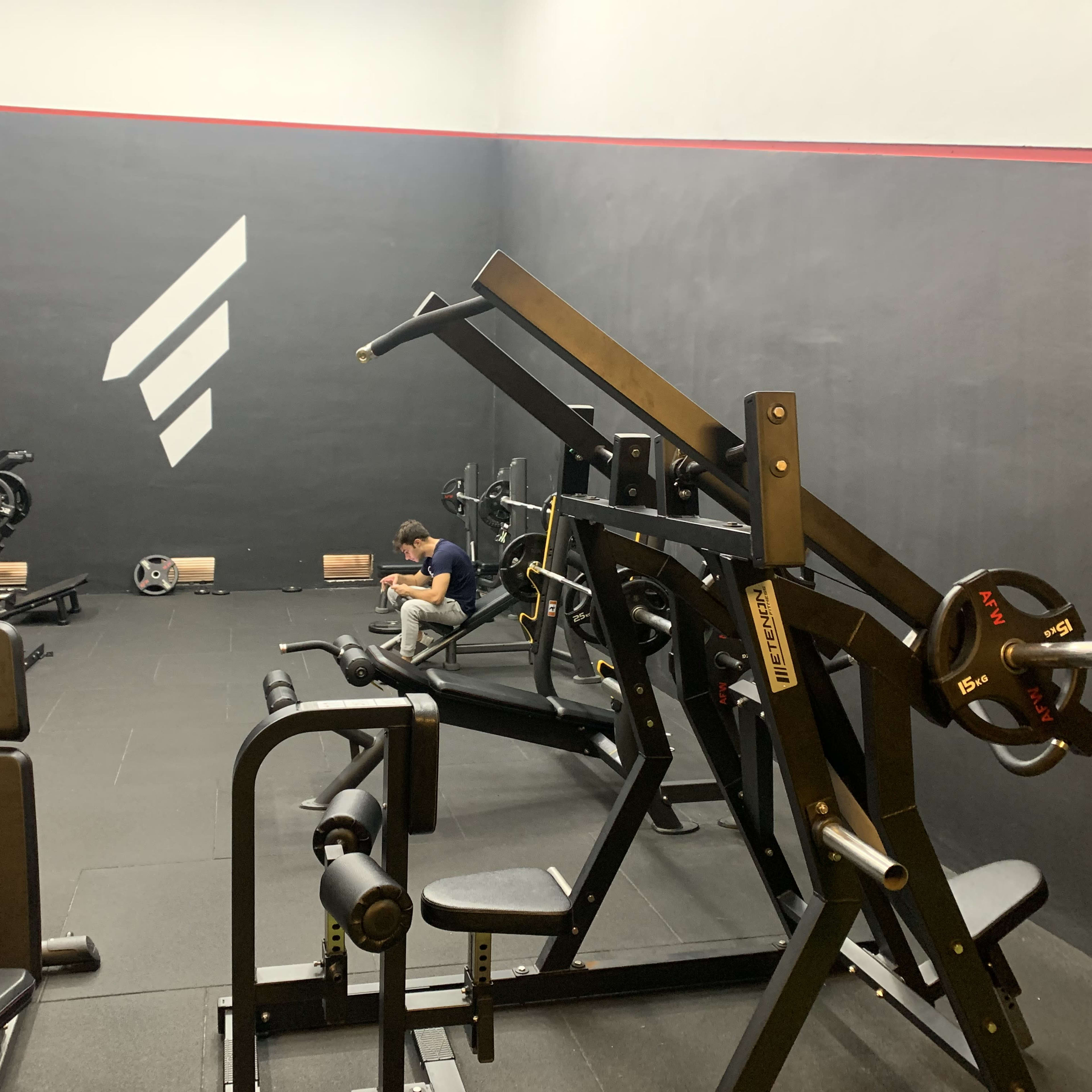 A small gym space features various weightlifting machines and a person seated near a wall, focused on their phone.