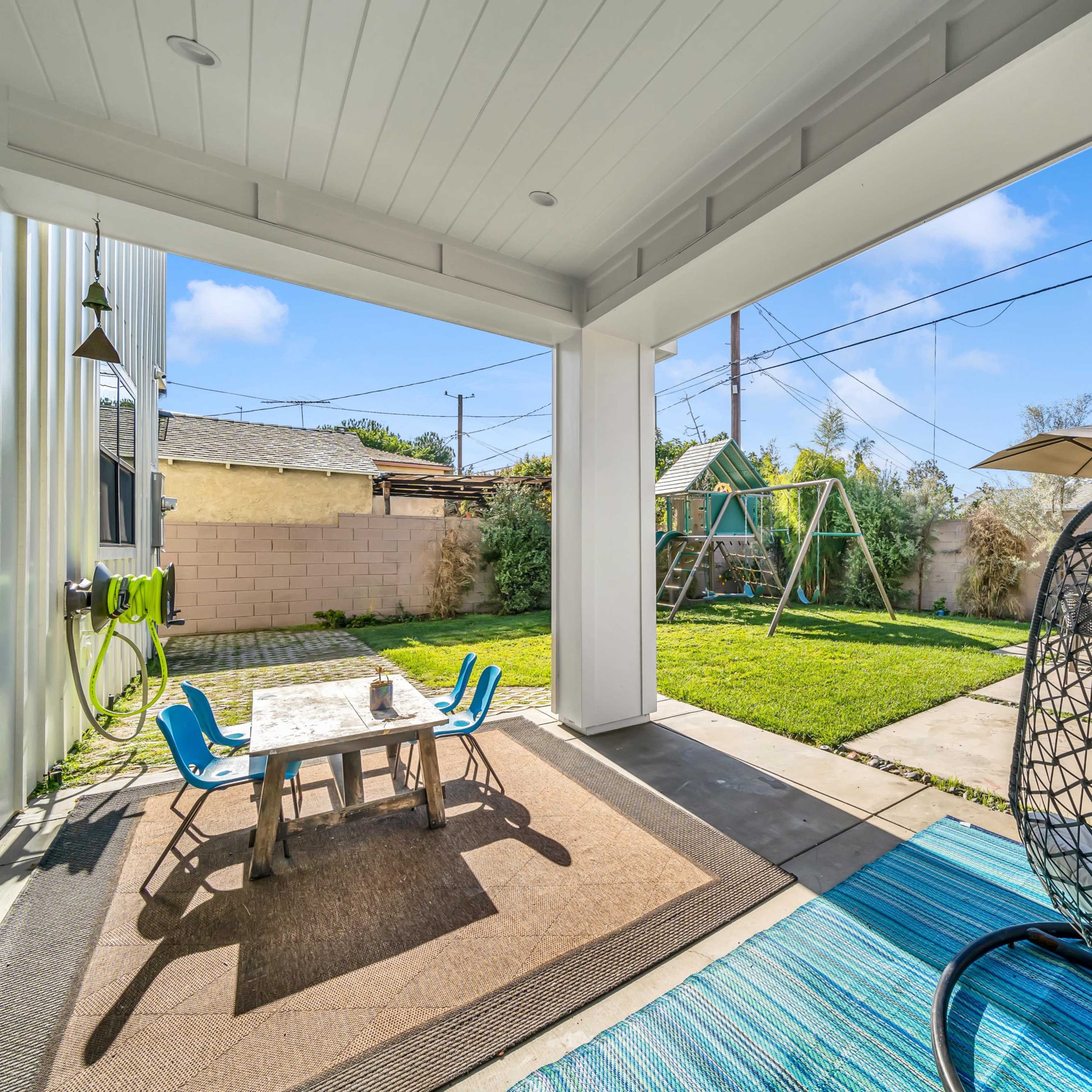 A backyard patio with a hanging chair, a small table and chairs, and a swing set visible in the background.