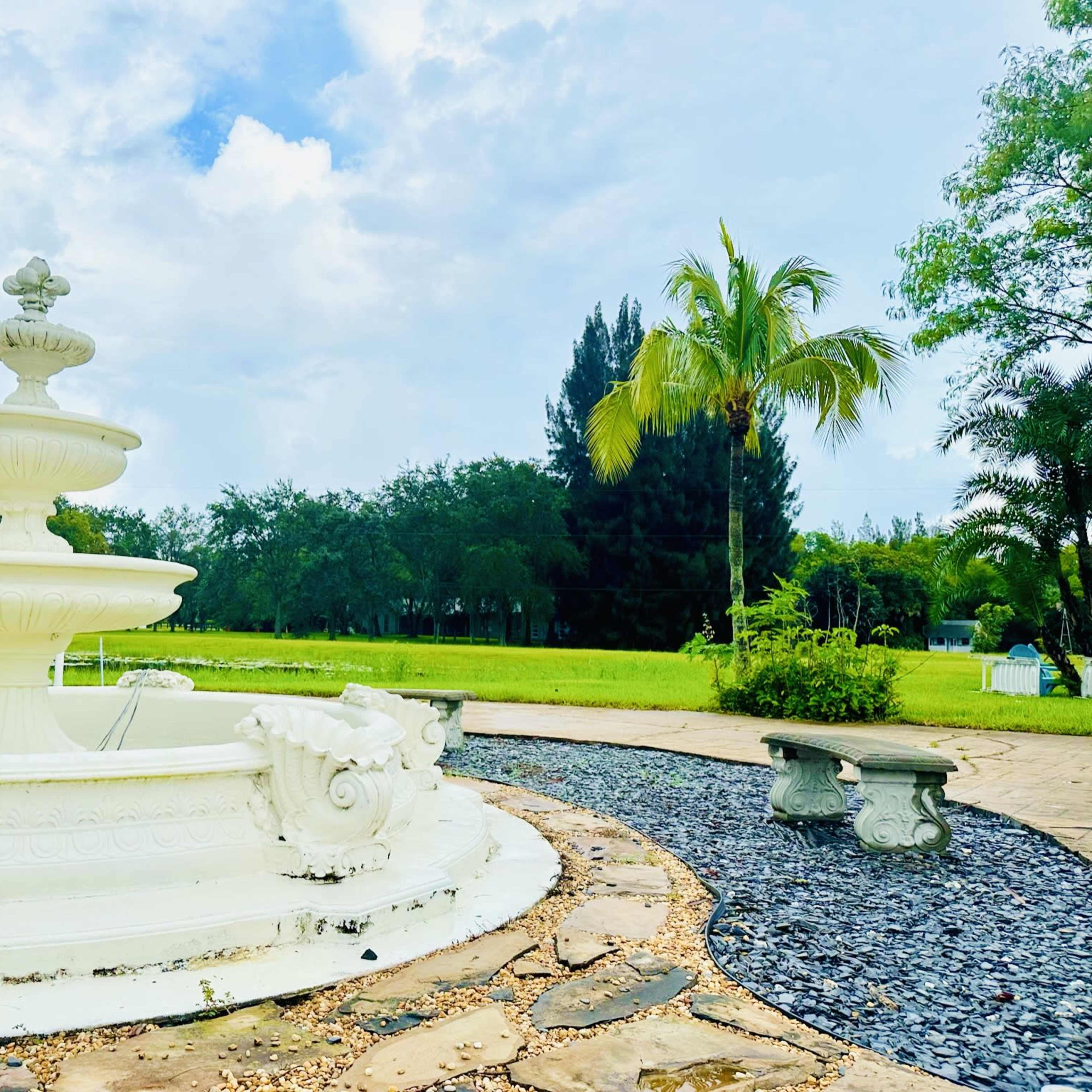 A white fountain stands in a landscaped area with stone paths and greenery under a partly cloudy sky.