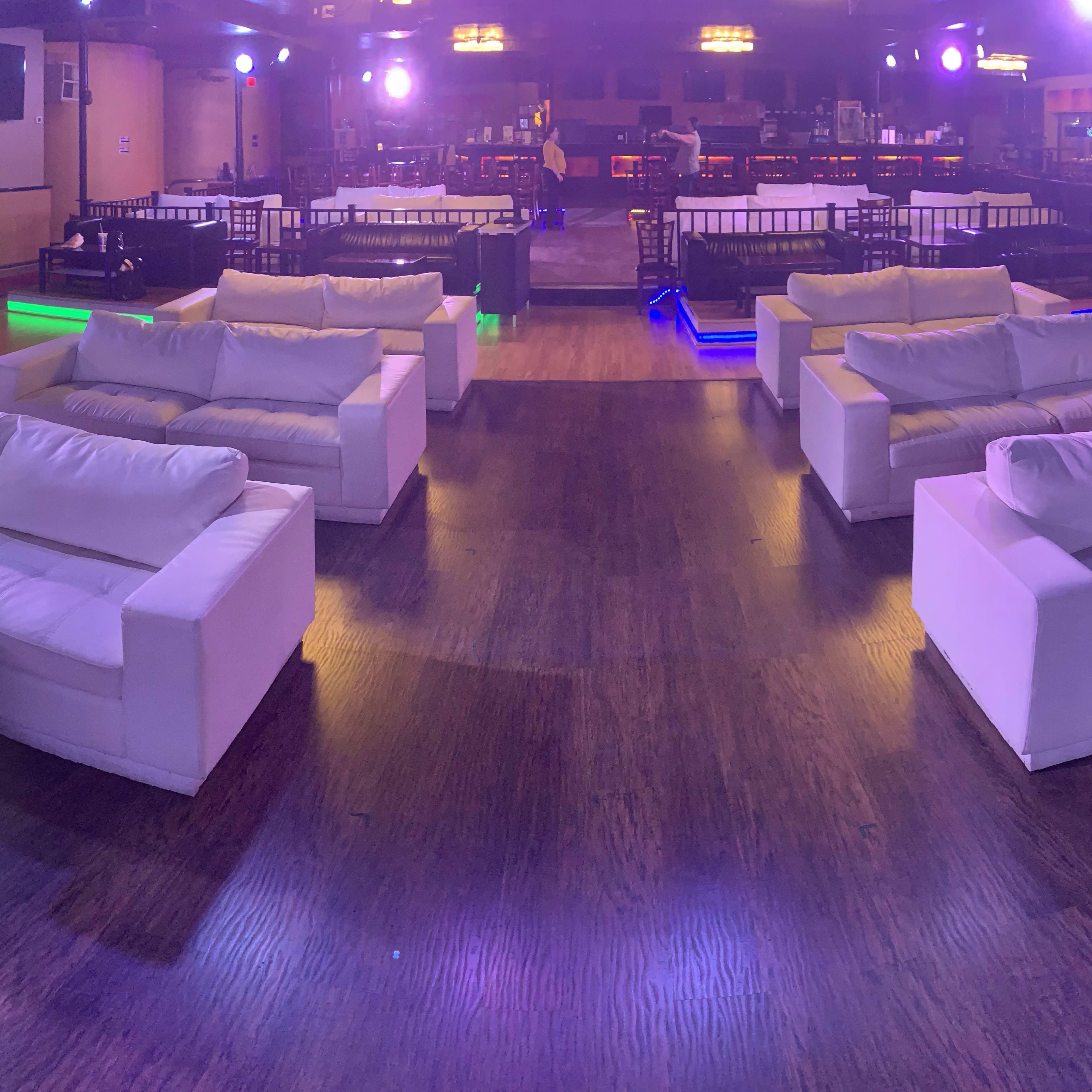 A spacious lounge area with white couches arranged around low tables, illuminated by colorful lights.