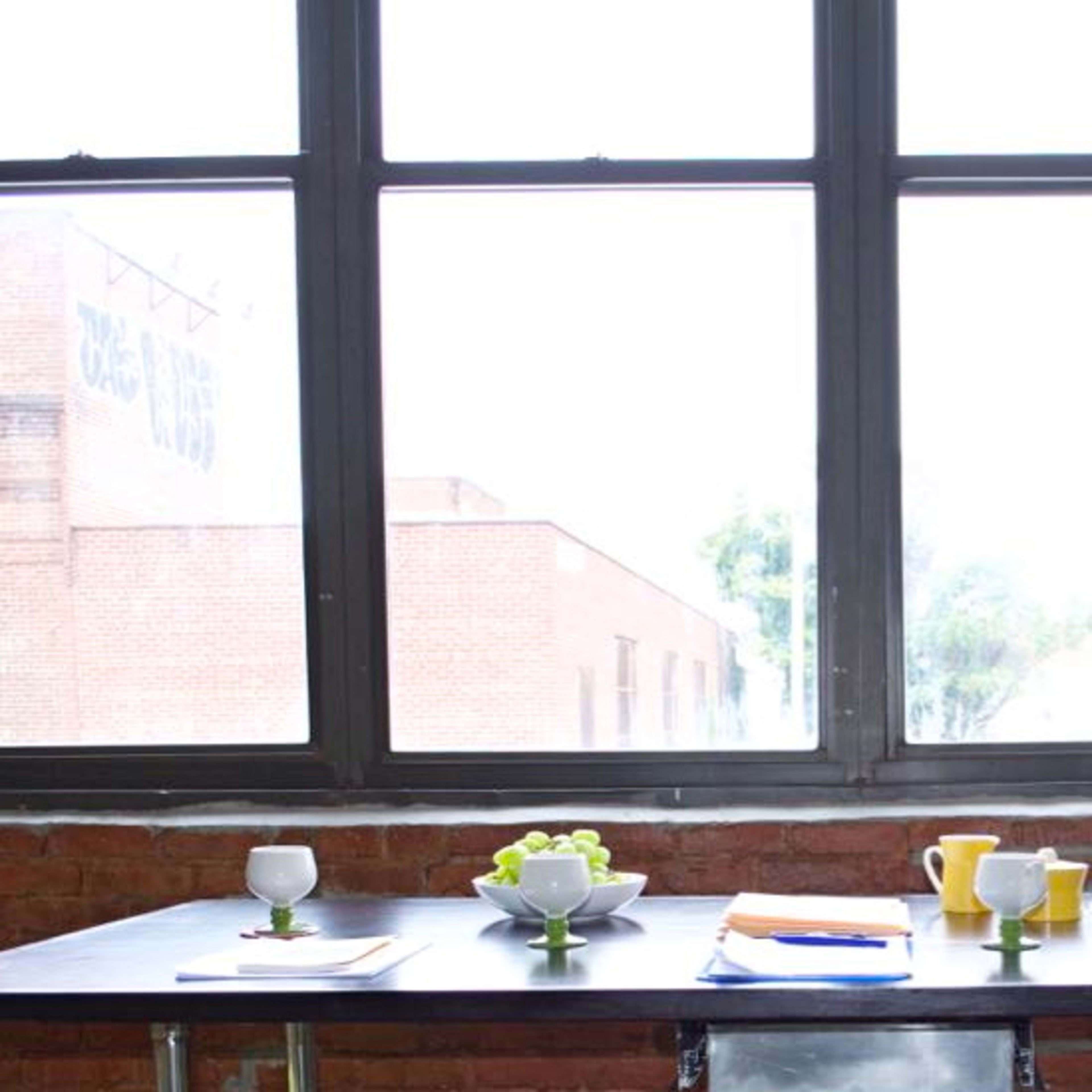 A dining table is set with dishes and drinks in front of large windows with a view of a brick wall outside.
