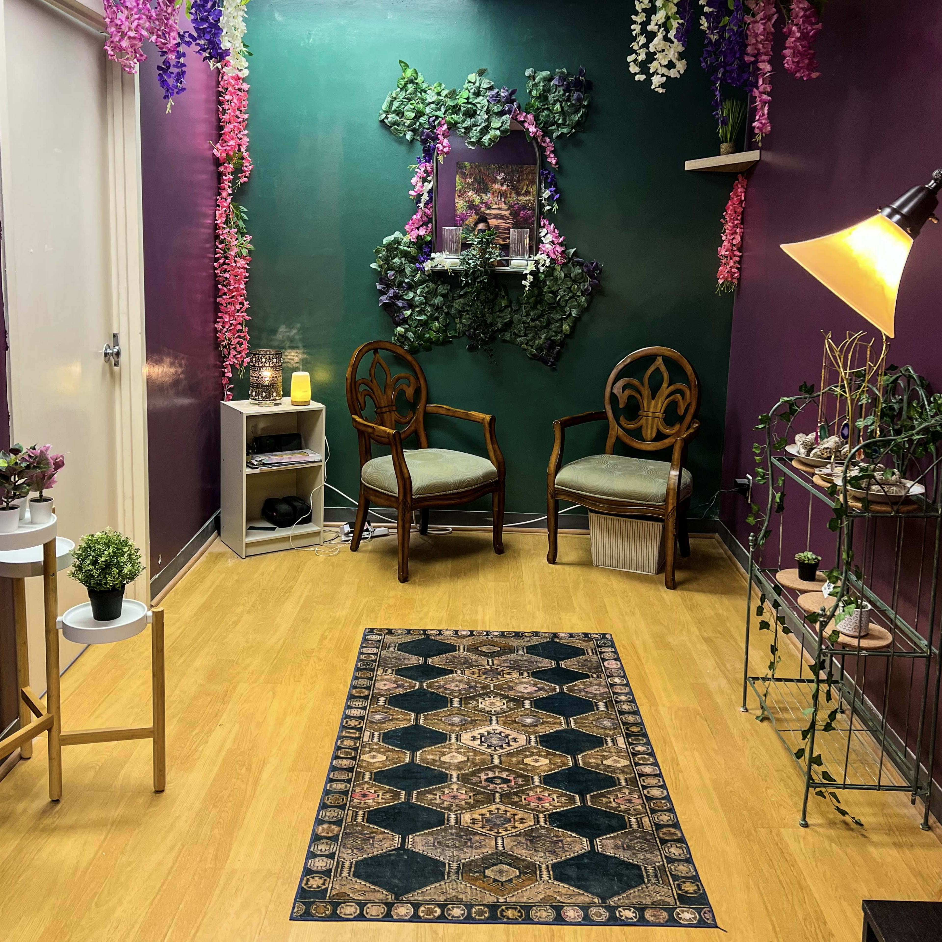 A cozy waiting area with two wooden chairs, green and purple walls adorned with faux plants, and a decorative rug on a wooden floor.