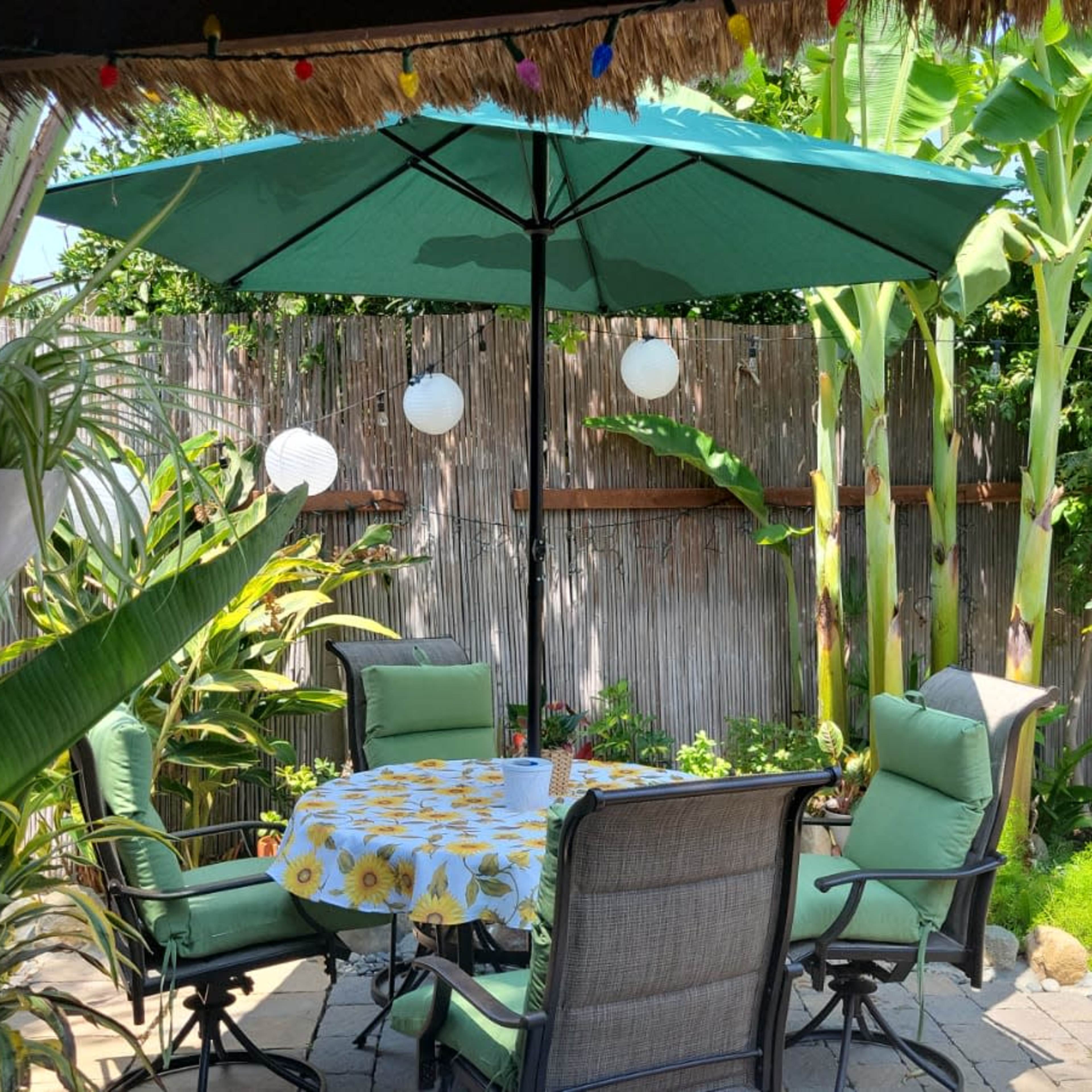 A shaded patio with a round table and four chairs surrounded by tropical plants and decorative lanterns.