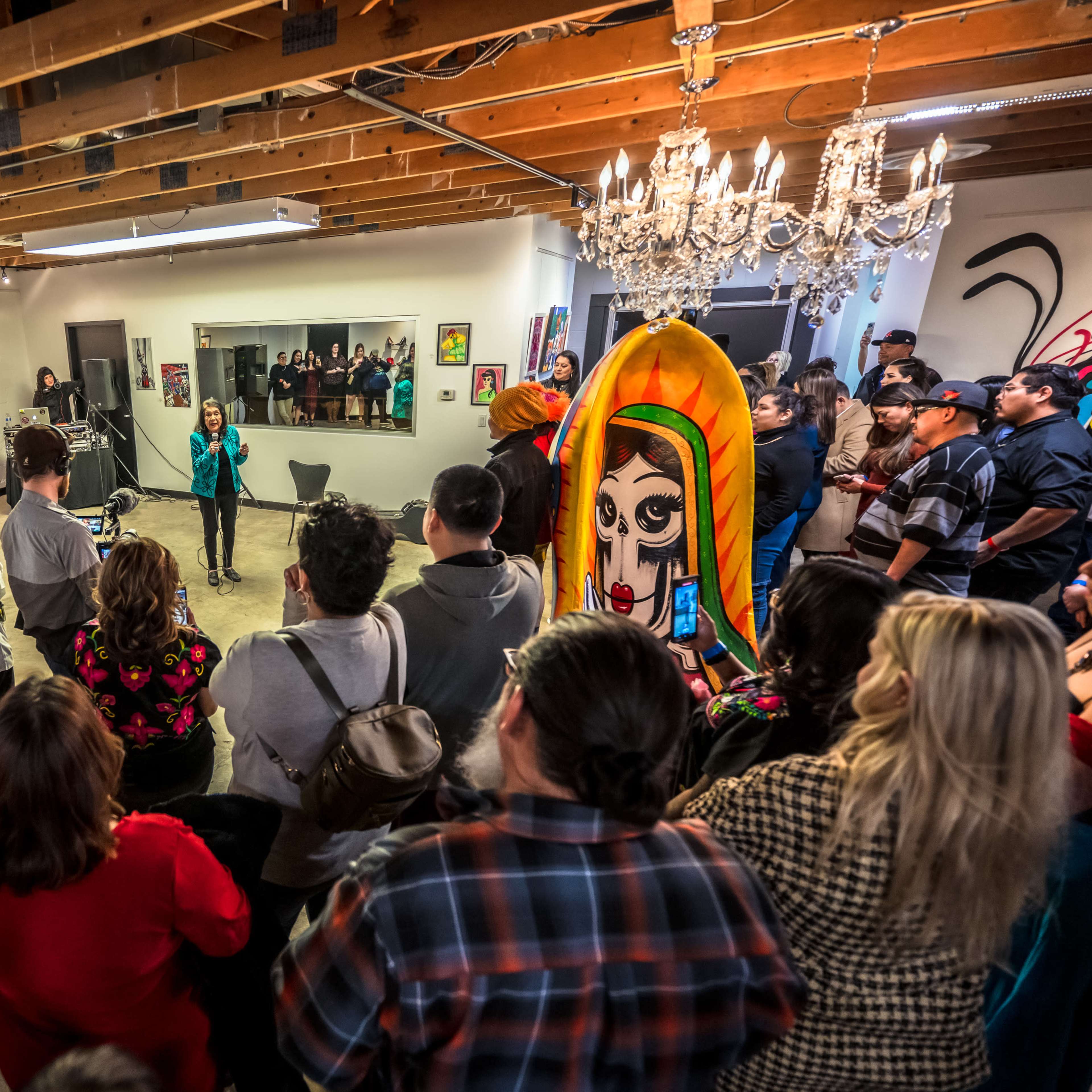 A diverse group of people gathers in an art gallery, attentively listening to a speaker in front of colorful artwork and a large, painted surfboard.