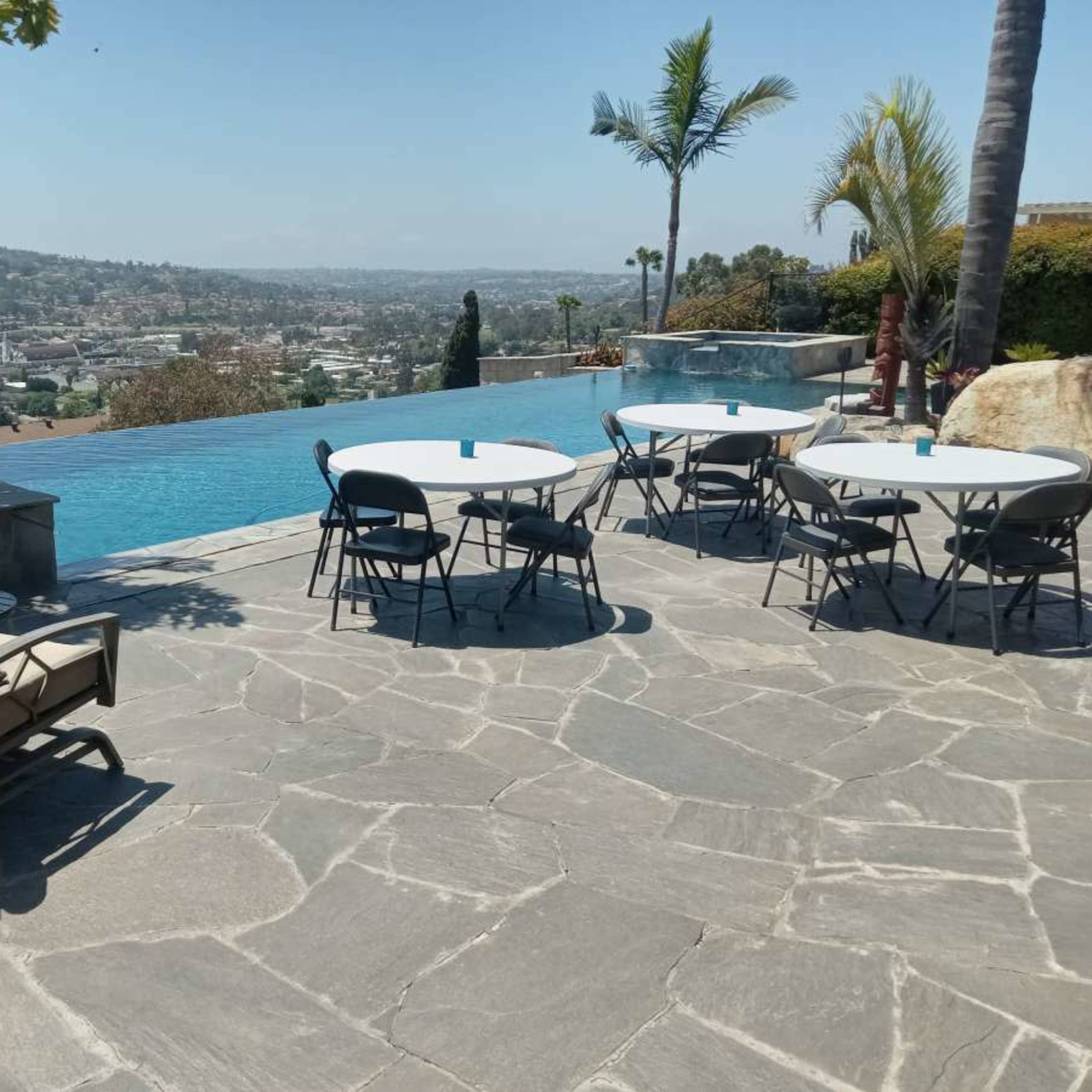 A patio area features several round tables and chairs beside a large infinity pool overlooking a cityscape.