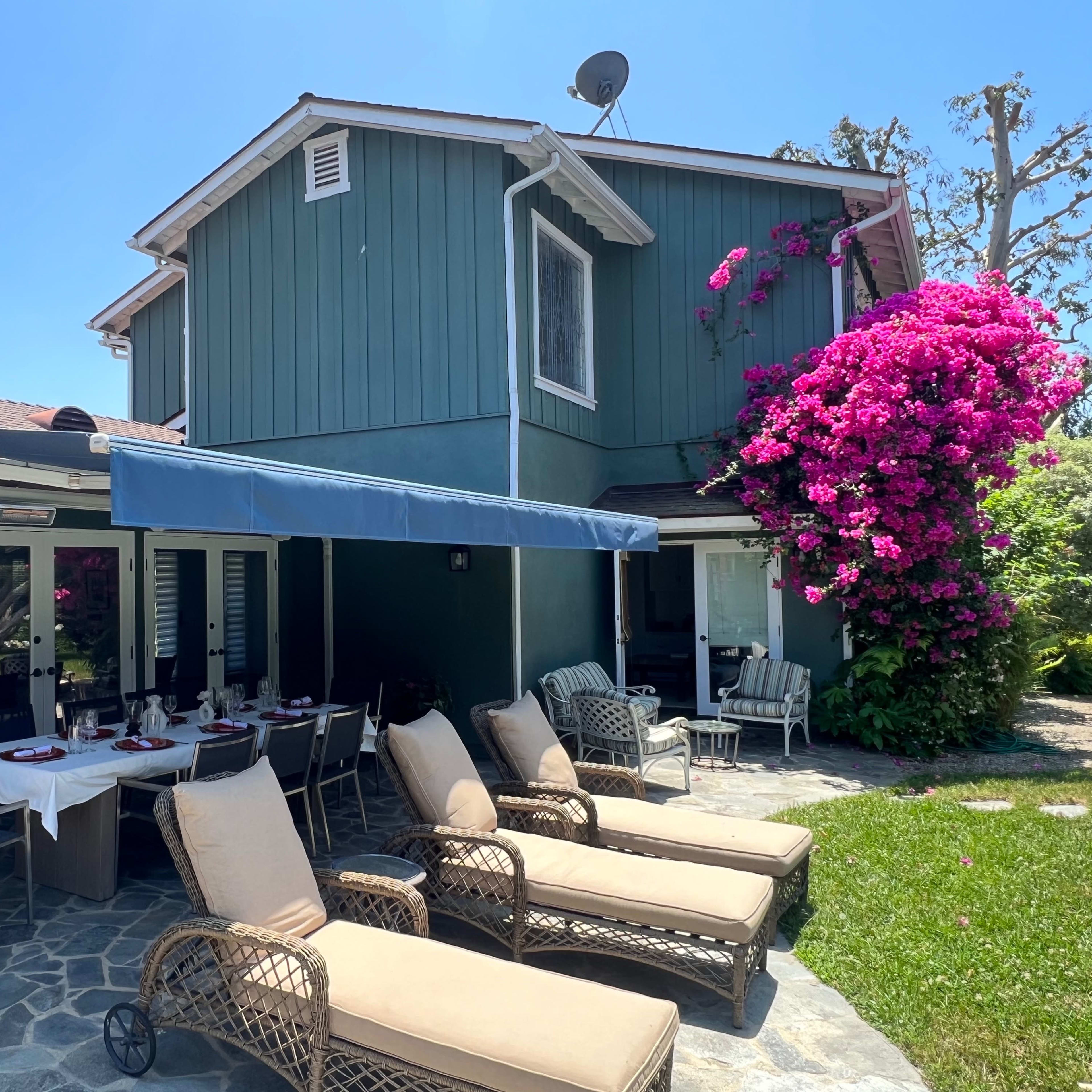 A patio with lounge chairs and a dining area is situated next to a green house adorned with blooming bougainvillea.