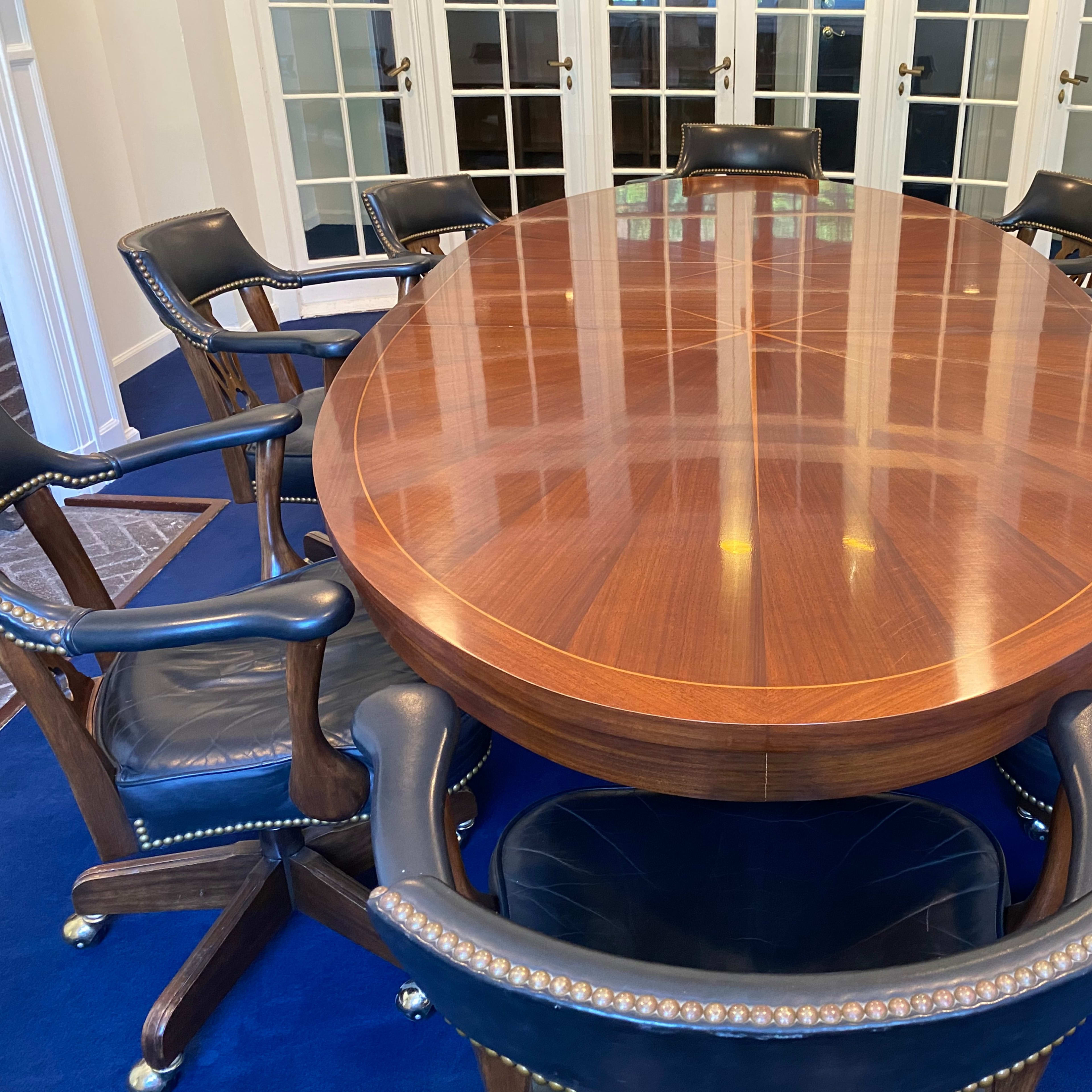 A large oval wooden conference table surrounded by eight chairs in a well-lit room with glass doors.