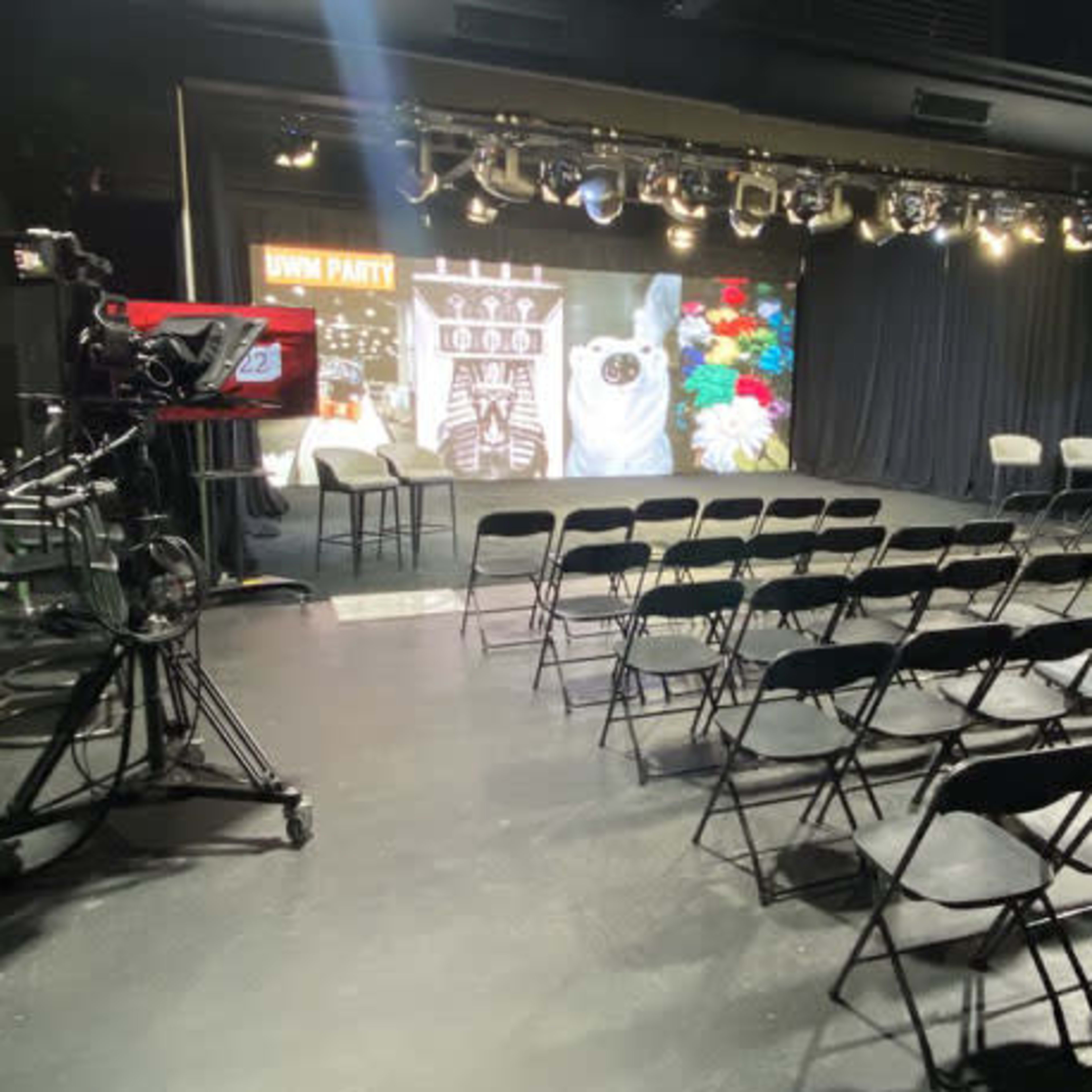 A television studio setup with multiple cameras, a green screen, and rows of black chairs facing a stage adorned with decorative elements.