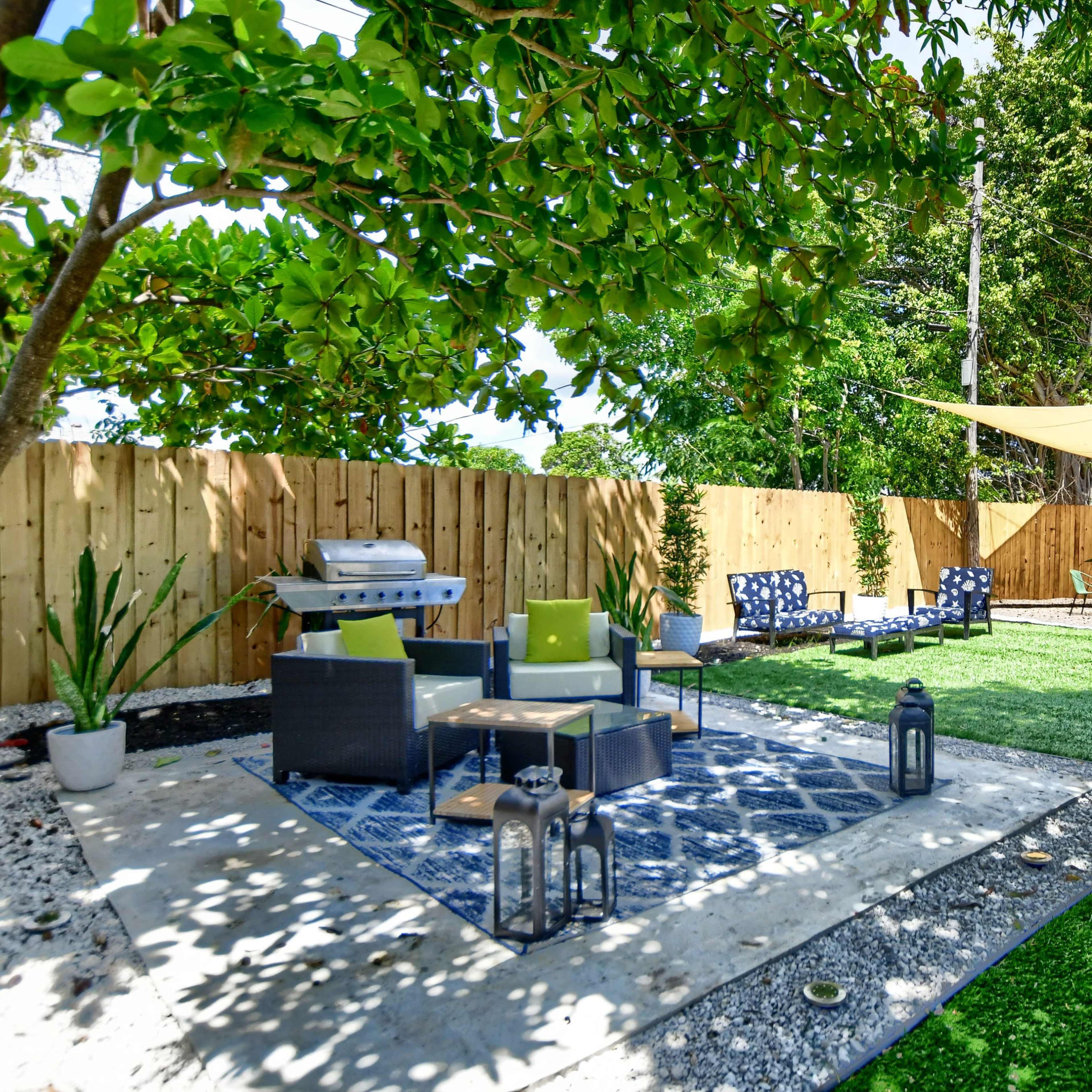 A landscaped outdoor seating area with a shaded canopy, comfortable chairs, a grill, and decorative plants surrounded by a wooden fence.