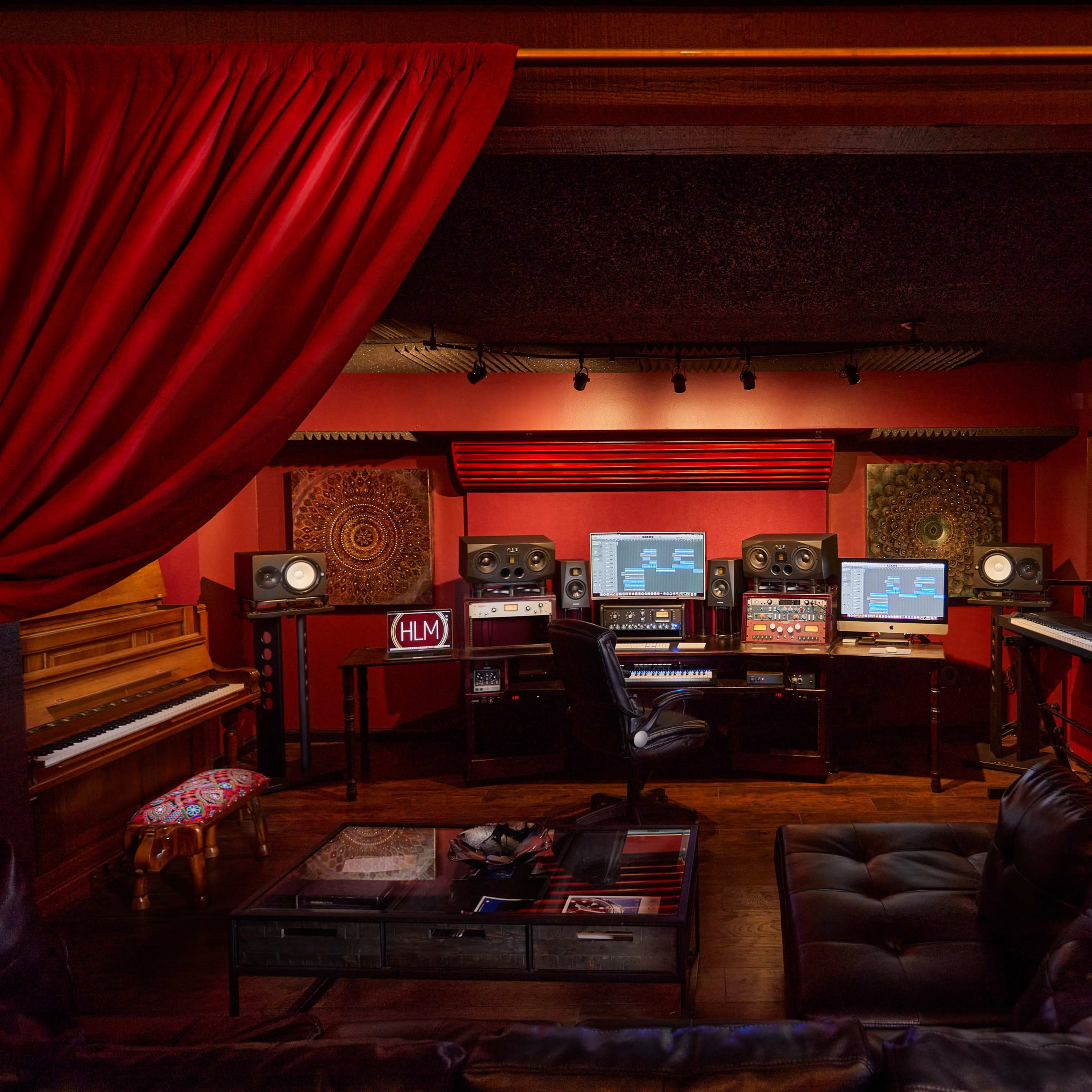 A recording studio featuring two computer monitors, audio equipment, a grand piano, and a black leather couch, all arranged within a room with red curtains and walls.