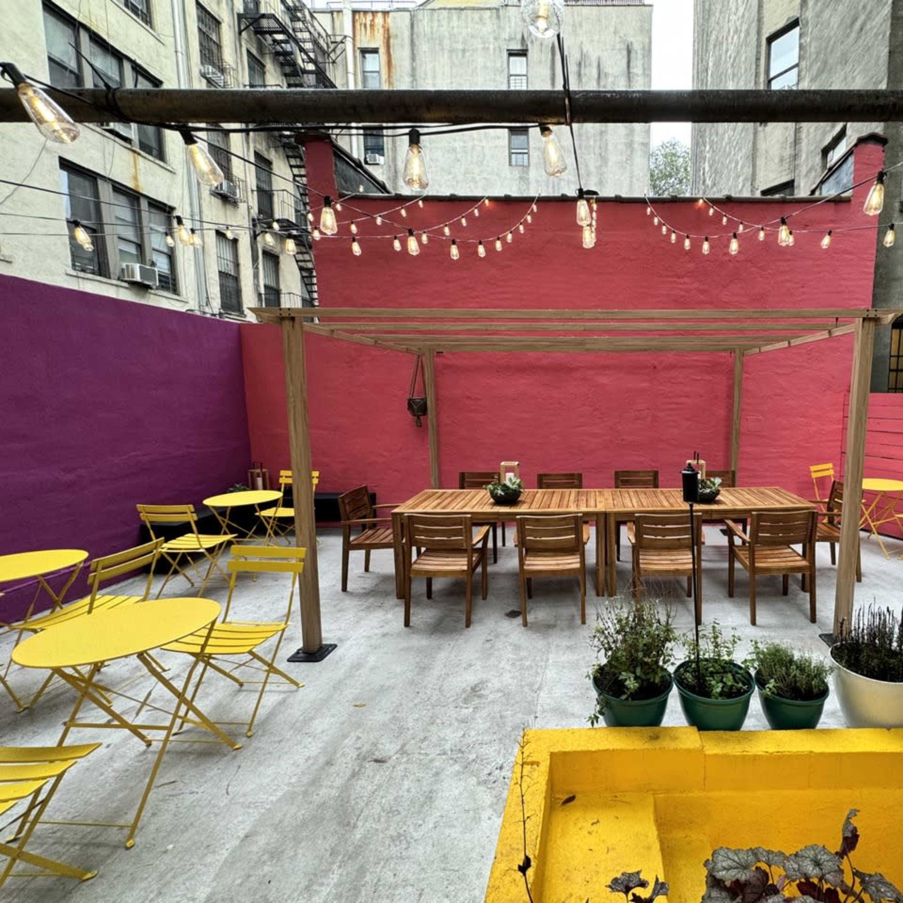 An outdoor patio space featuring yellow metal chairs, wooden tables, and a pink wall, illuminated by string lights.