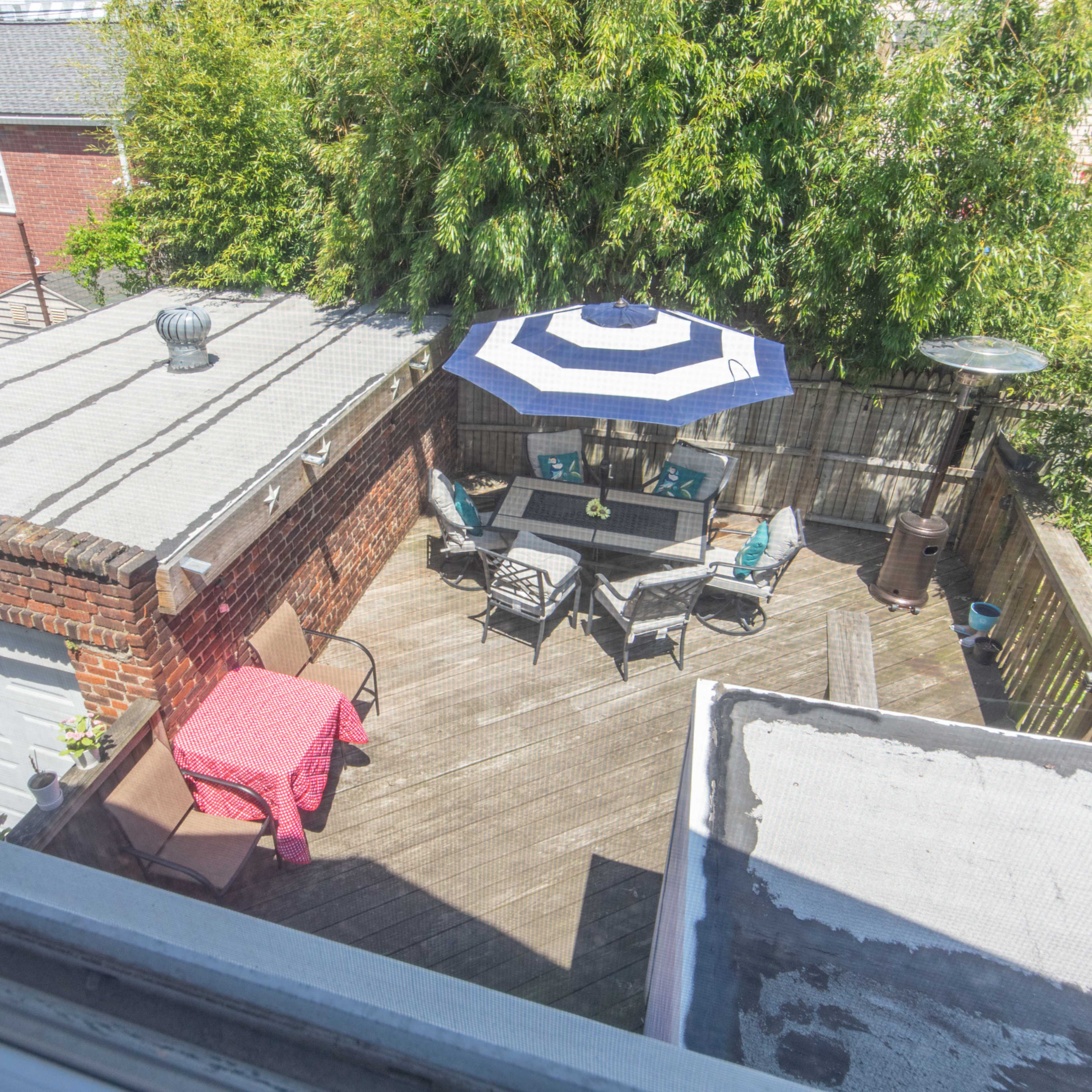 A rooftop patio with a table and chairs, featuring a large umbrella, surrounded by greenery and adjacent buildings.