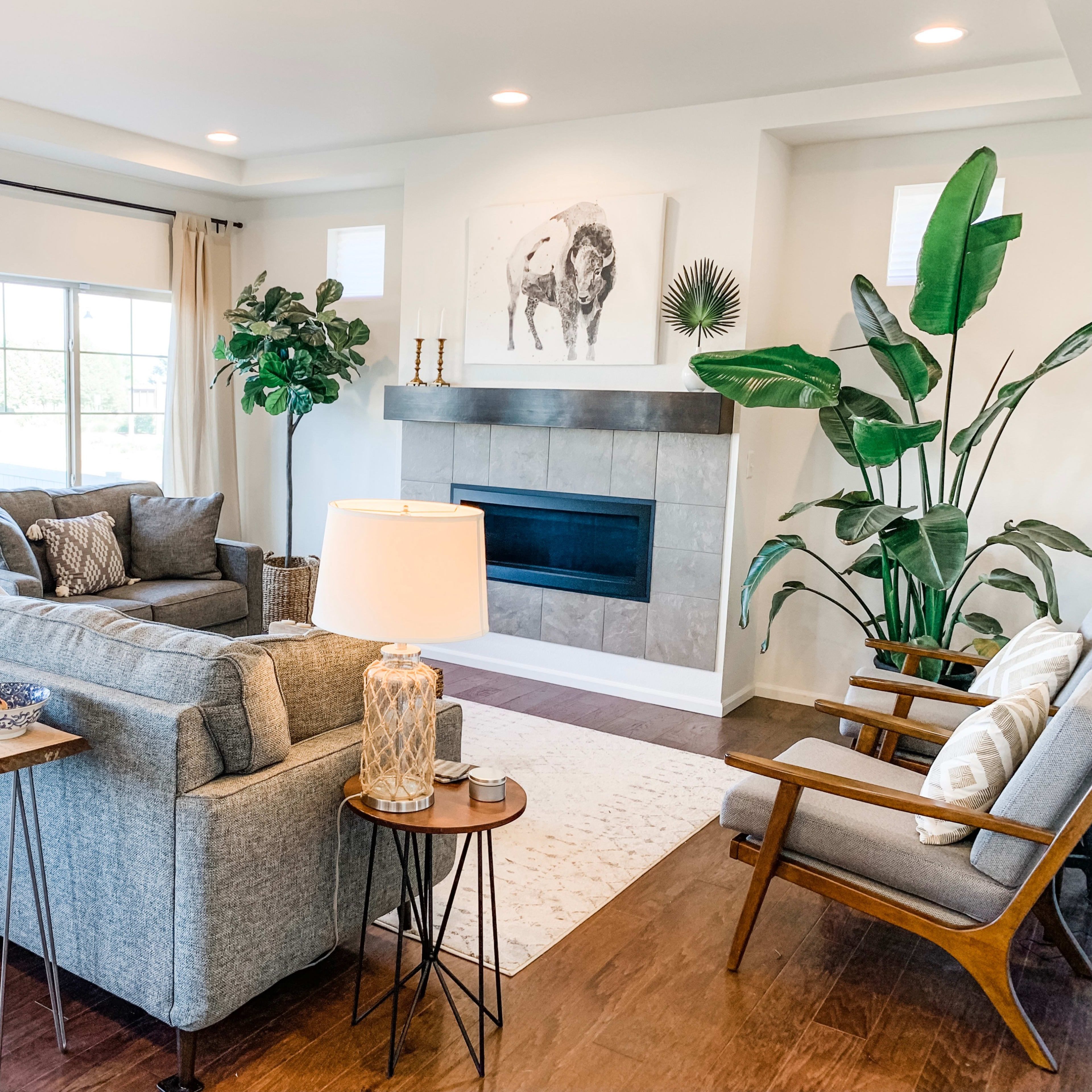 A cozy living room features a gray sofa, wooden chairs, a fireplace, and large windows with natural light.