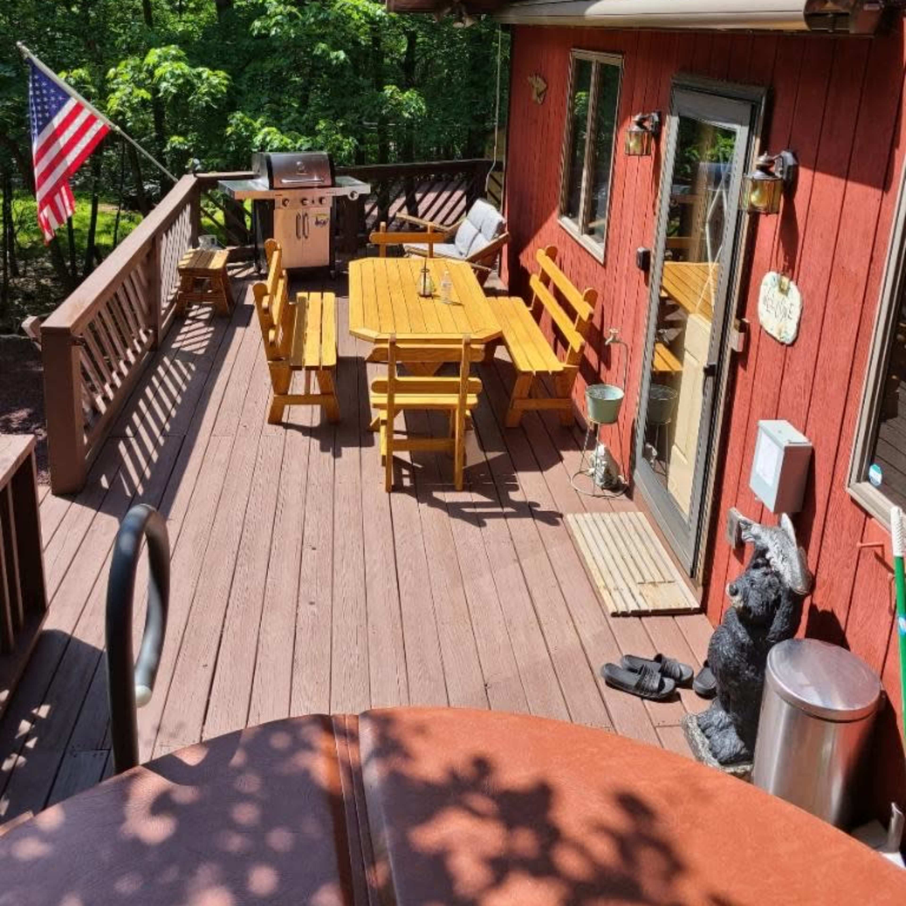 A wooden deck with multiple picnic tables, a grill, and an American flag, surrounded by lush green trees.