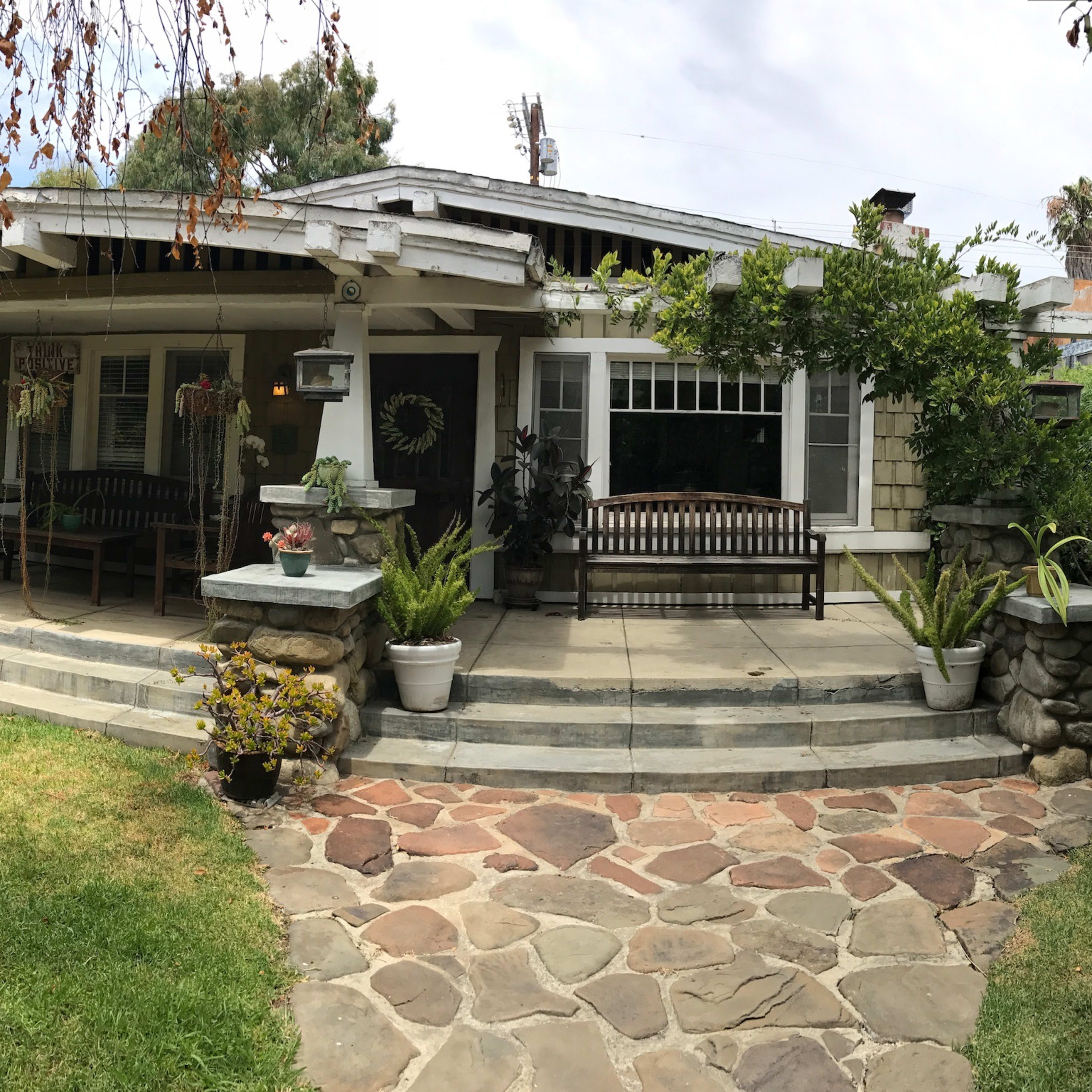 A single-story house with a wide porch, surrounded by a landscaped yard featuring stone pathways and various plants.