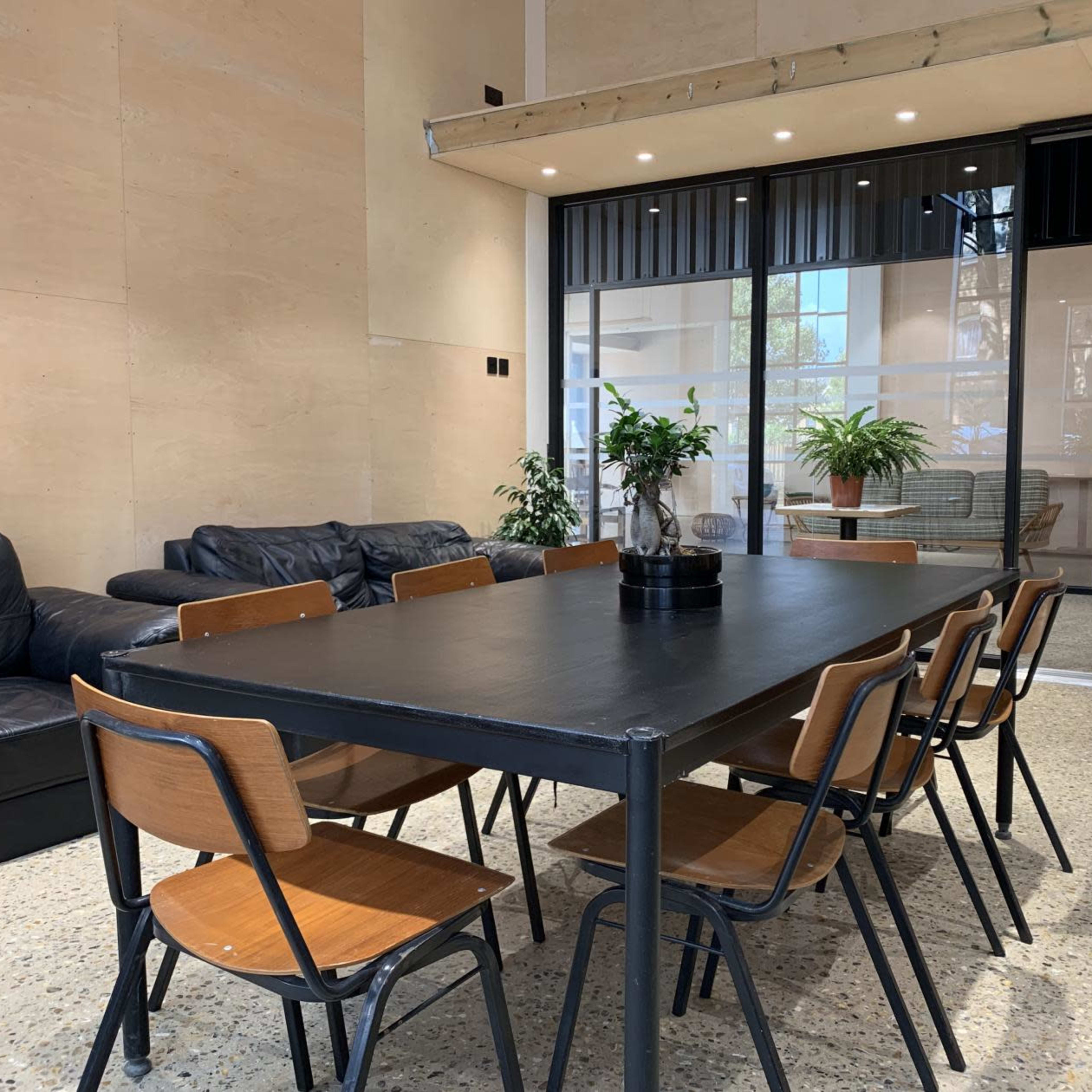A modern meeting room with a long black table surrounded by wooden chairs, and a couch area in the background.