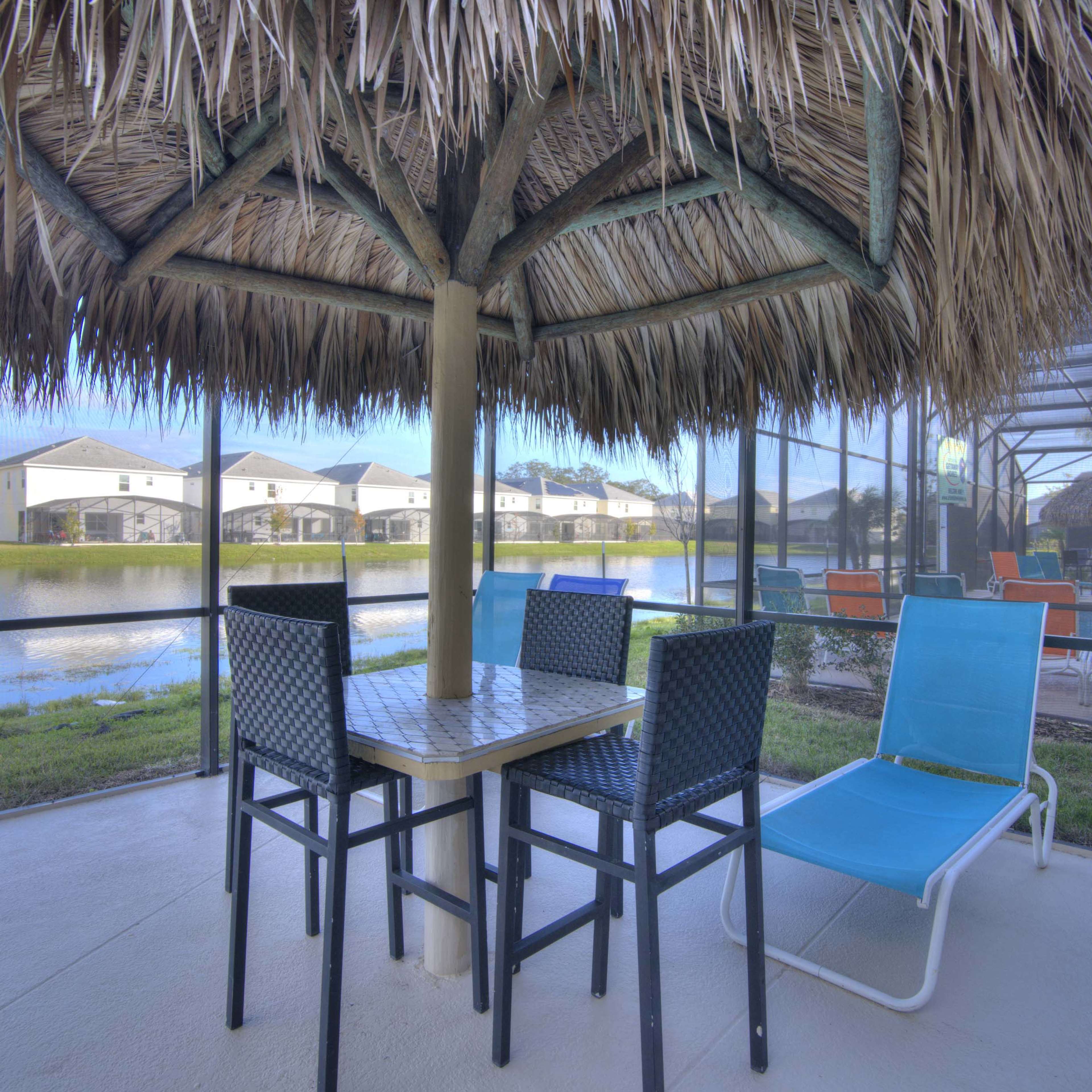 A thatched-roof patio gazebo with a table and chairs overlooks a calm pond surrounded by houses.