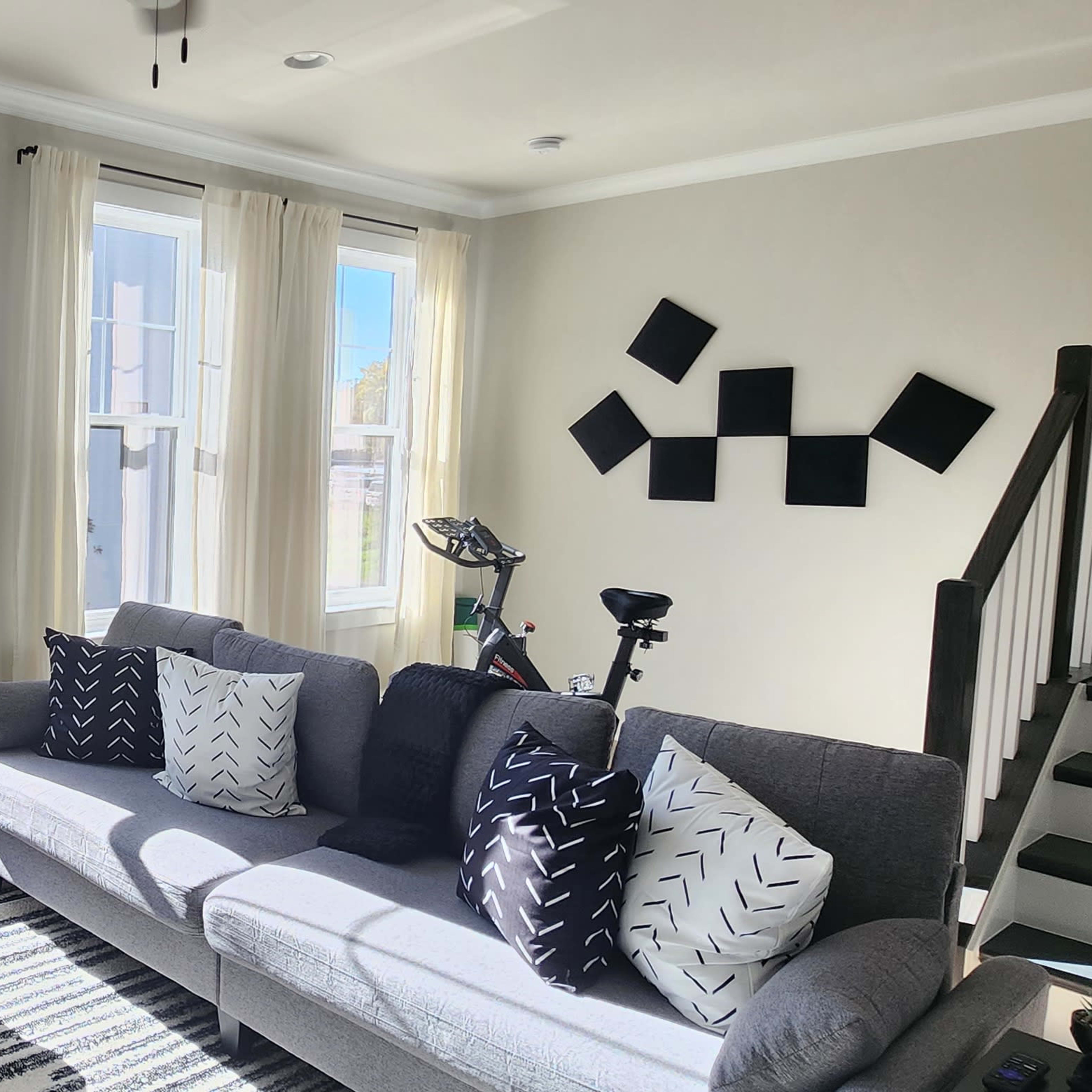 A bright living room with large windows, a gray sofa adorned with black and white pillows, an ottoman, a striped rug, and a staircase in the background.