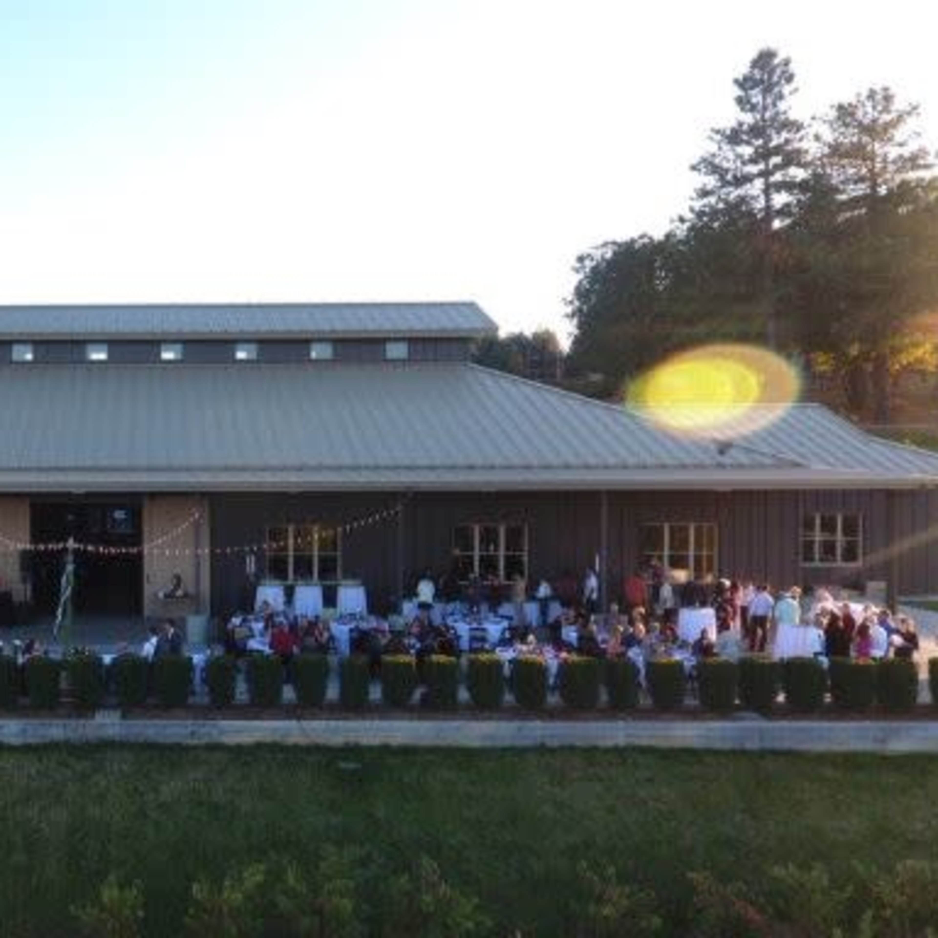 A large gathering of people is seated at tables arranged outside a metal building, with decorations and greenery lining the area.