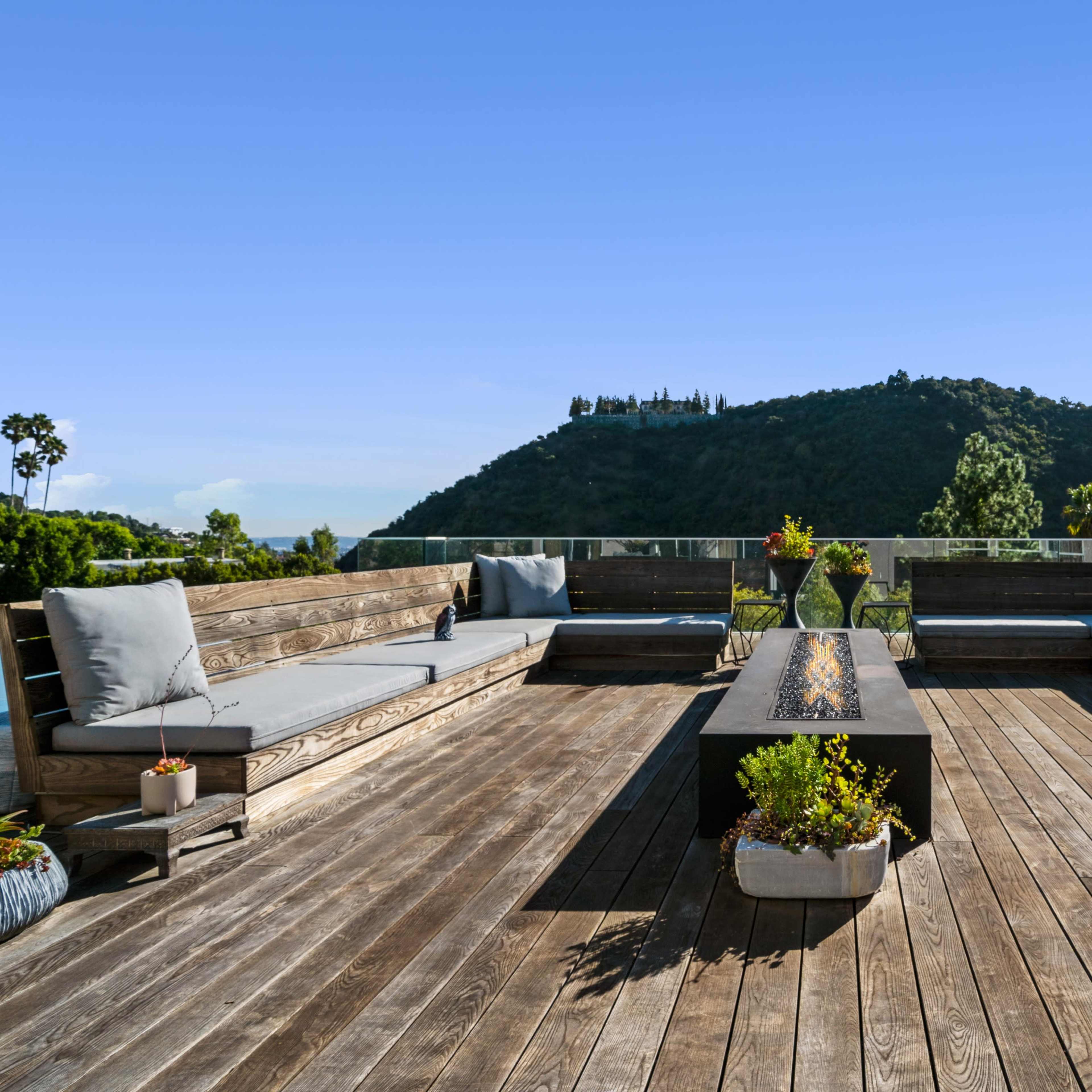 A spacious wooden deck features built-in seating and a central planter, set against a backdrop of lush hills.