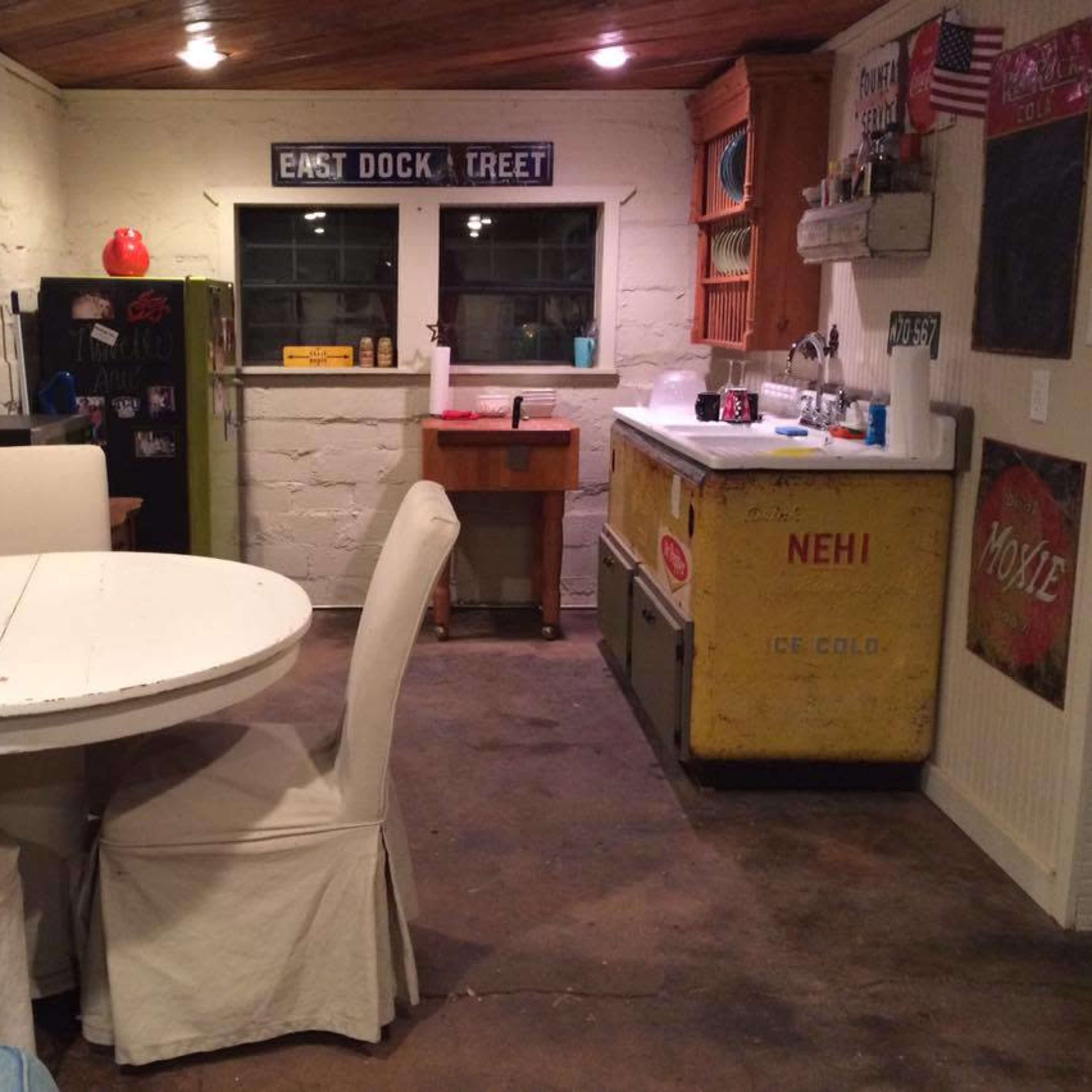 A rustic kitchen area with a round white table, four upholstered chairs, a yellow vintage ice cooler, and a window overlooking a small workspace.