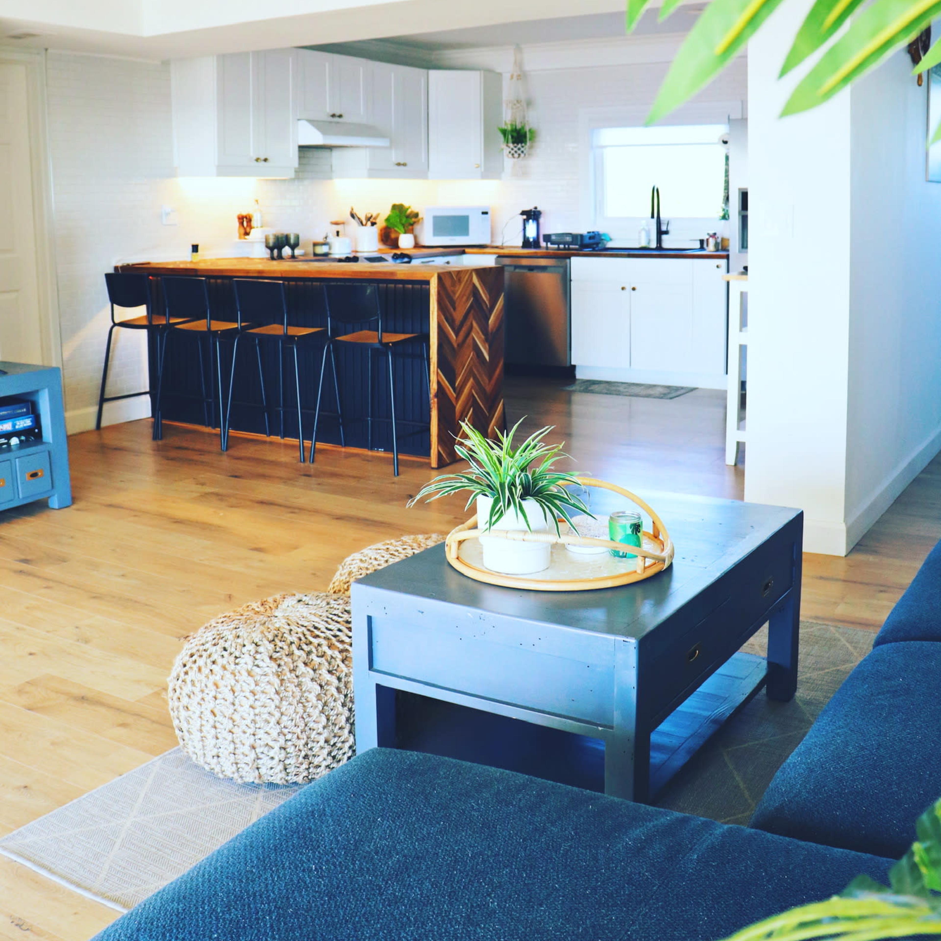 A modern living space with a kitchen in the background, featuring a dining area with bar stools, a coffee table, and a decorative plant.