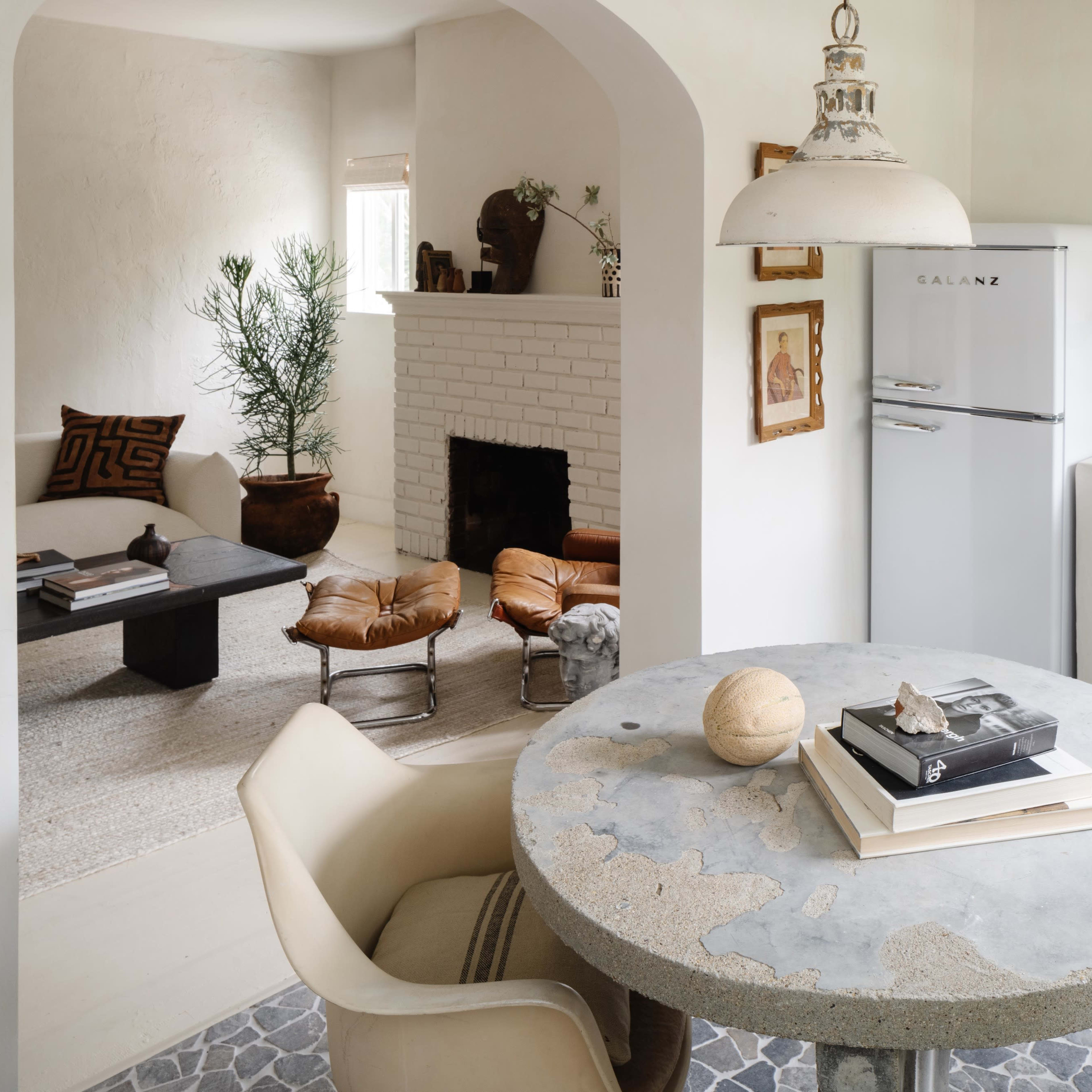 A minimalist kitchen and living area featuring a round concrete table with books on it, a retro-style refrigerator, and a cozy seating area with a fireplace.
