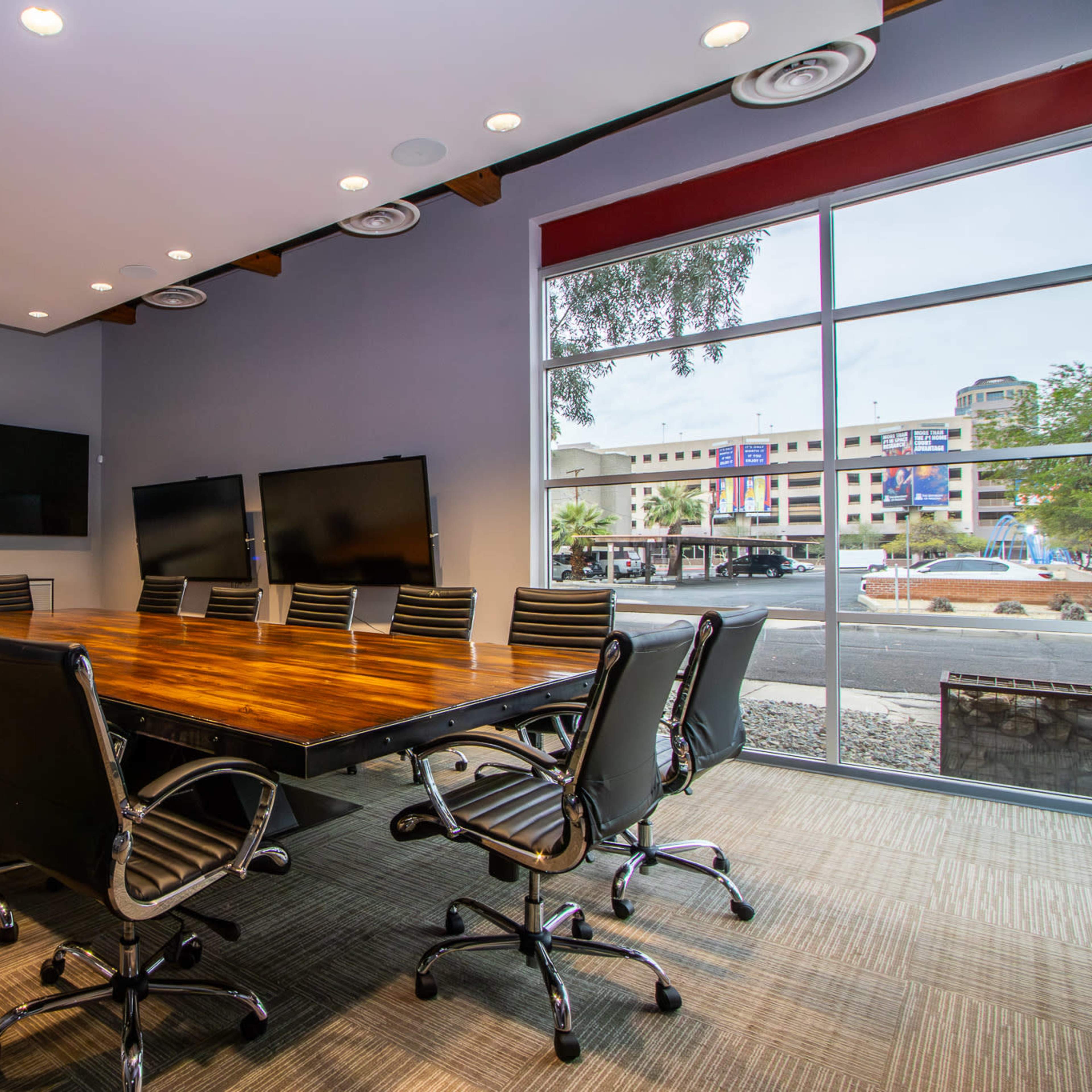 A conference room features a long wooden table surrounded by black leather chairs and multiple large screens on the walls.