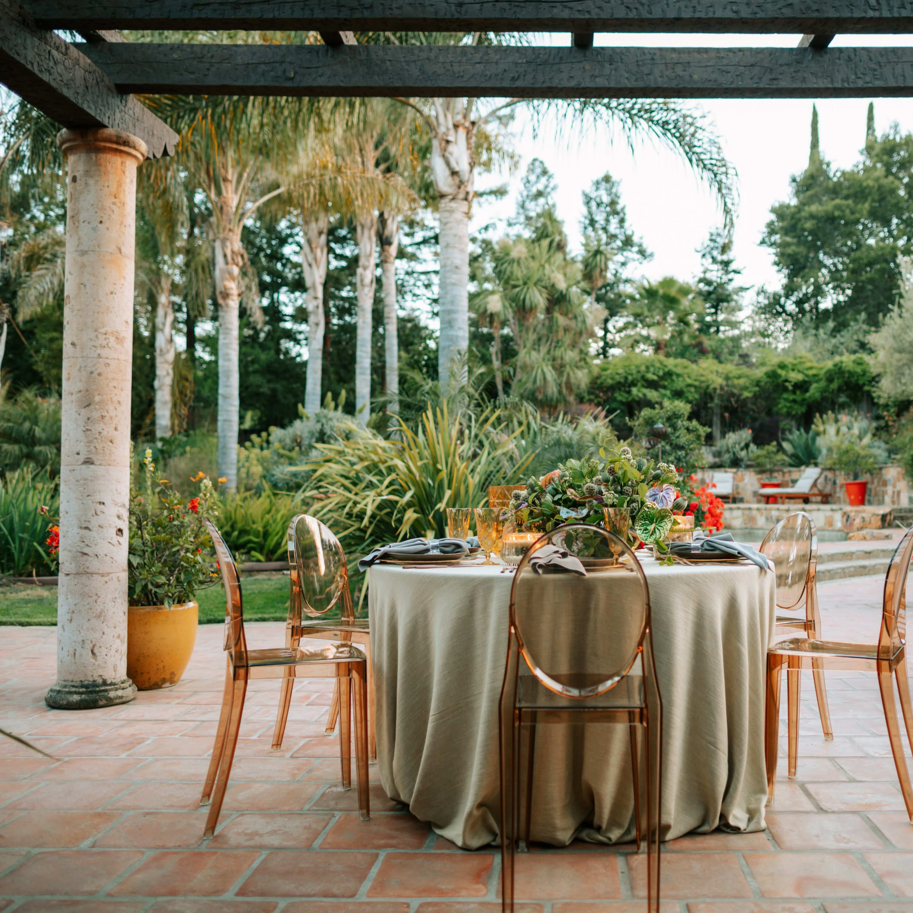 A circular table set for dining is placed under a pergola surrounded by lush greenery and potted plants.
