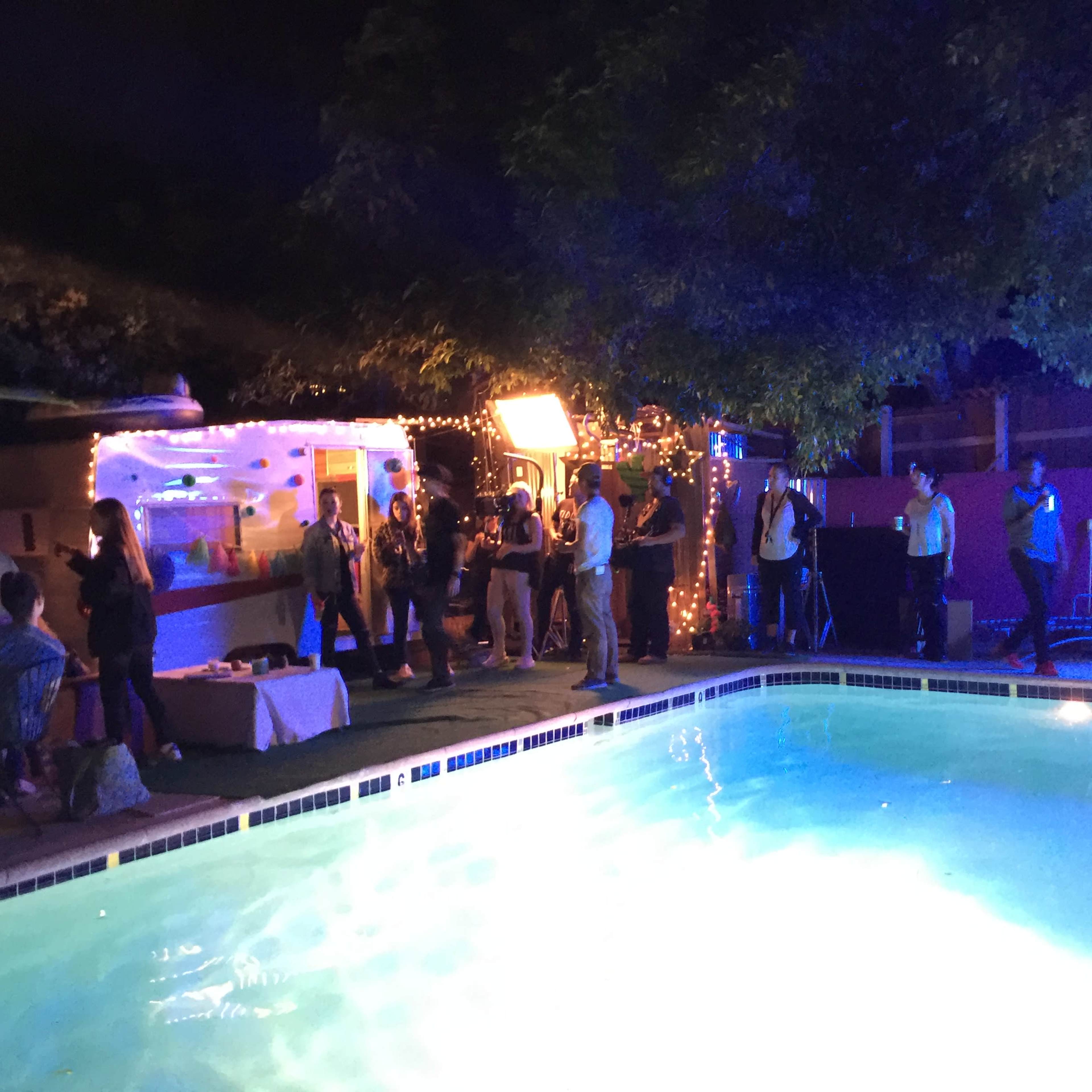 A group of people gathers around a poolside area illuminated by colorful lights during a night event.