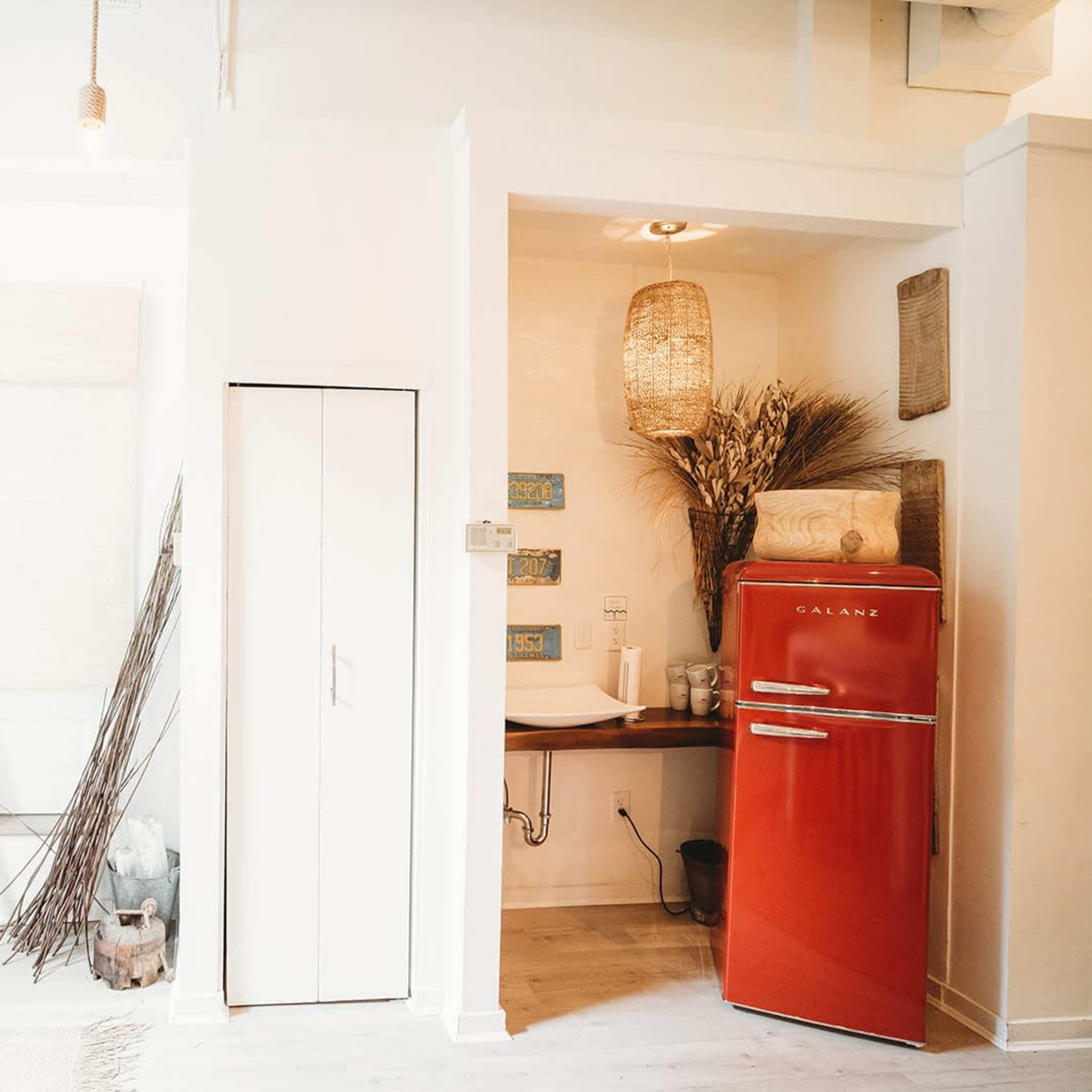 A bright interior space featuring a vintage red refrigerator, a small sink area with woven decor, and light-colored walls with natural elements.