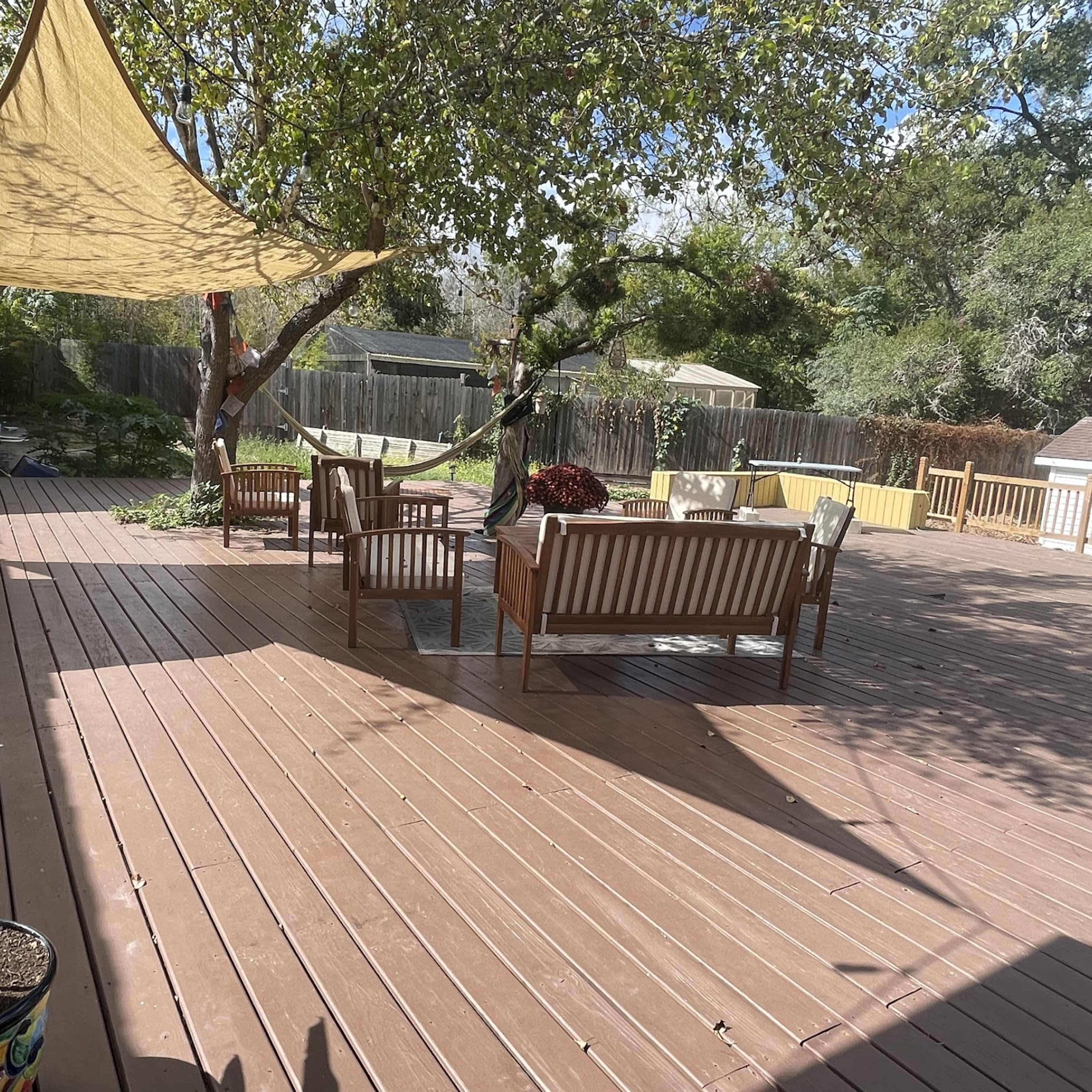 A wooden deck features several seating arrangements and a shade sail above, surrounded by trees and a storage shed.