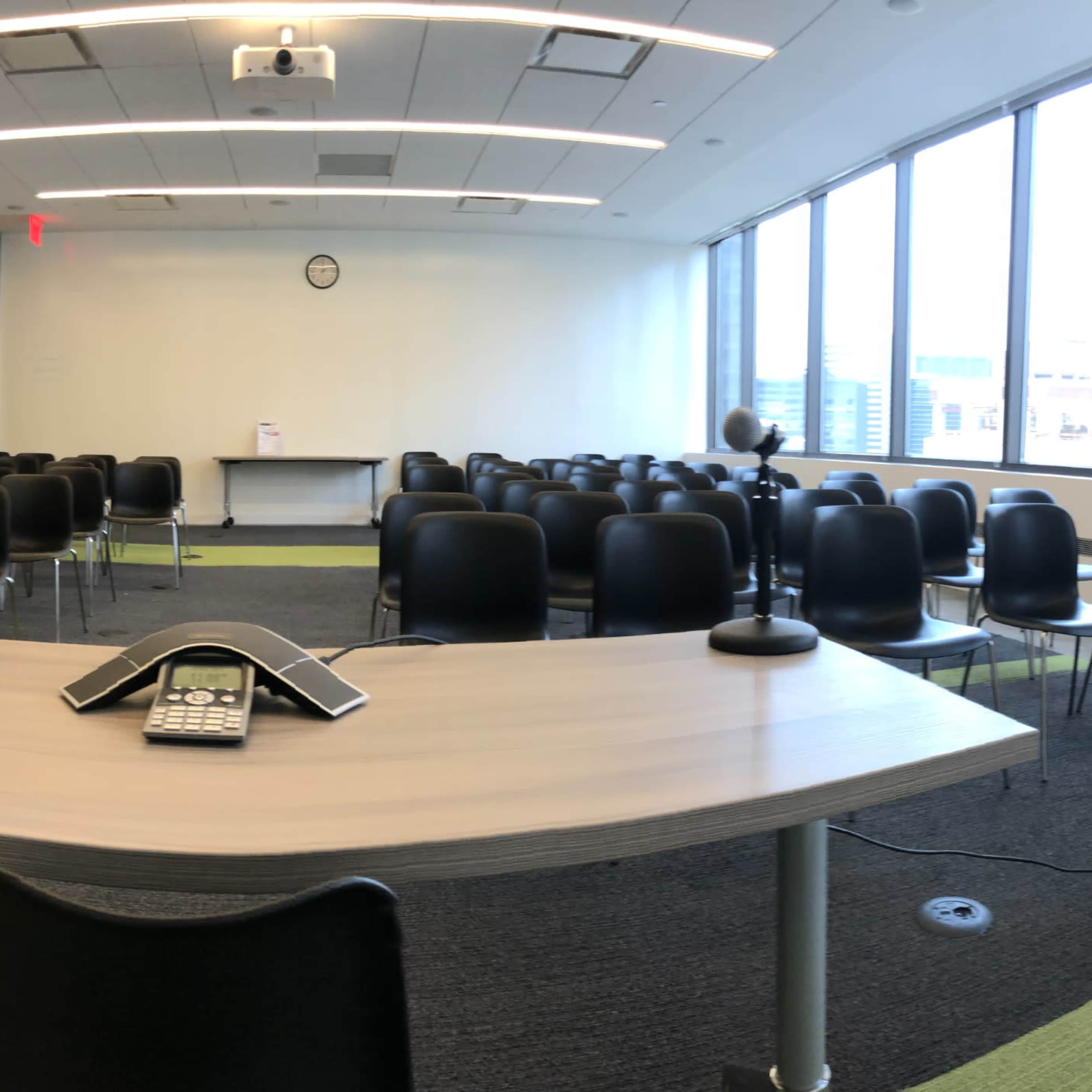 A conference room features a long table with a phone at the center and rows of black chairs set up for a gathering.