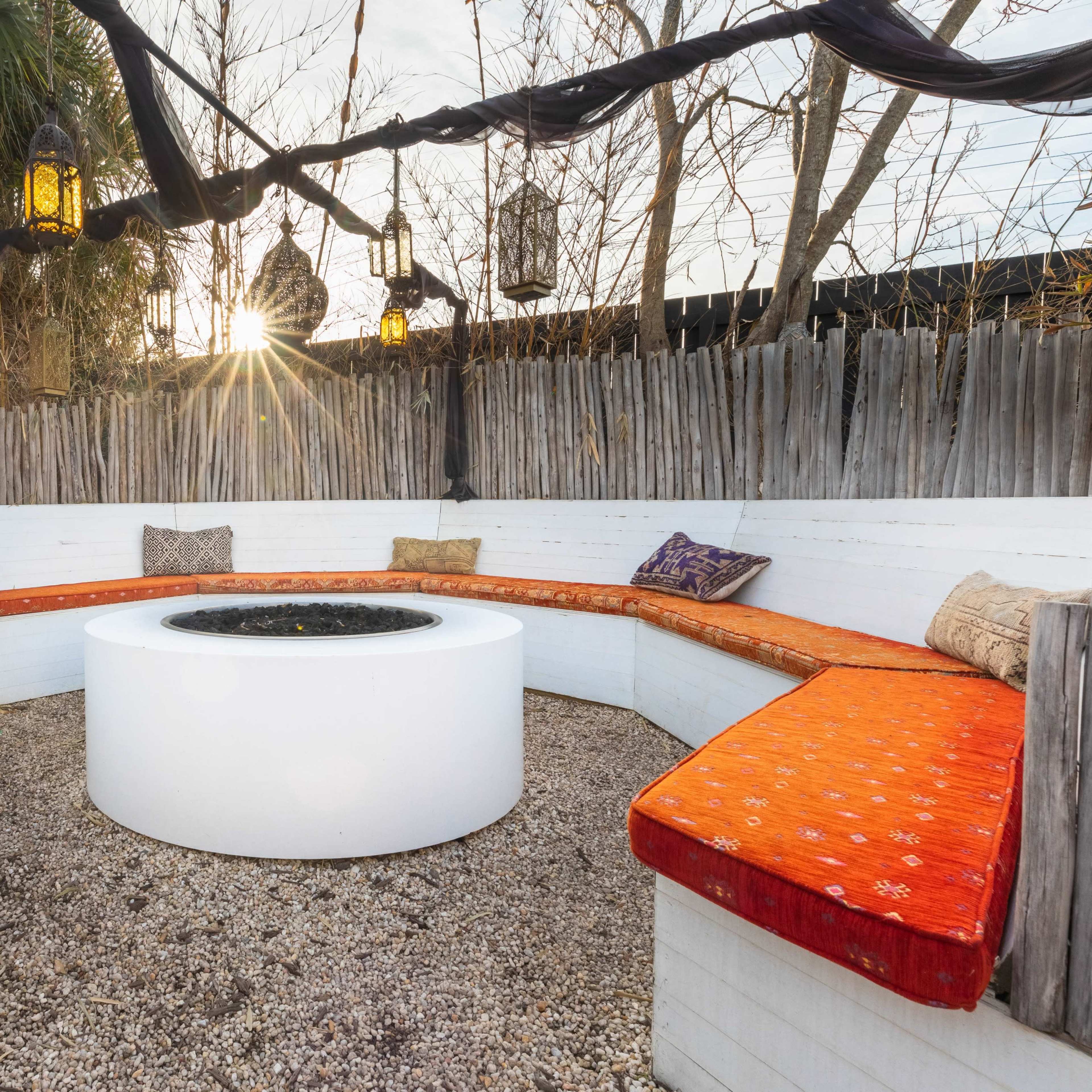 A circular seating area with orange cushions surrounds a central fire pit, set against a backdrop of wooden fencing and hanging lanterns, with a sunset in the background.