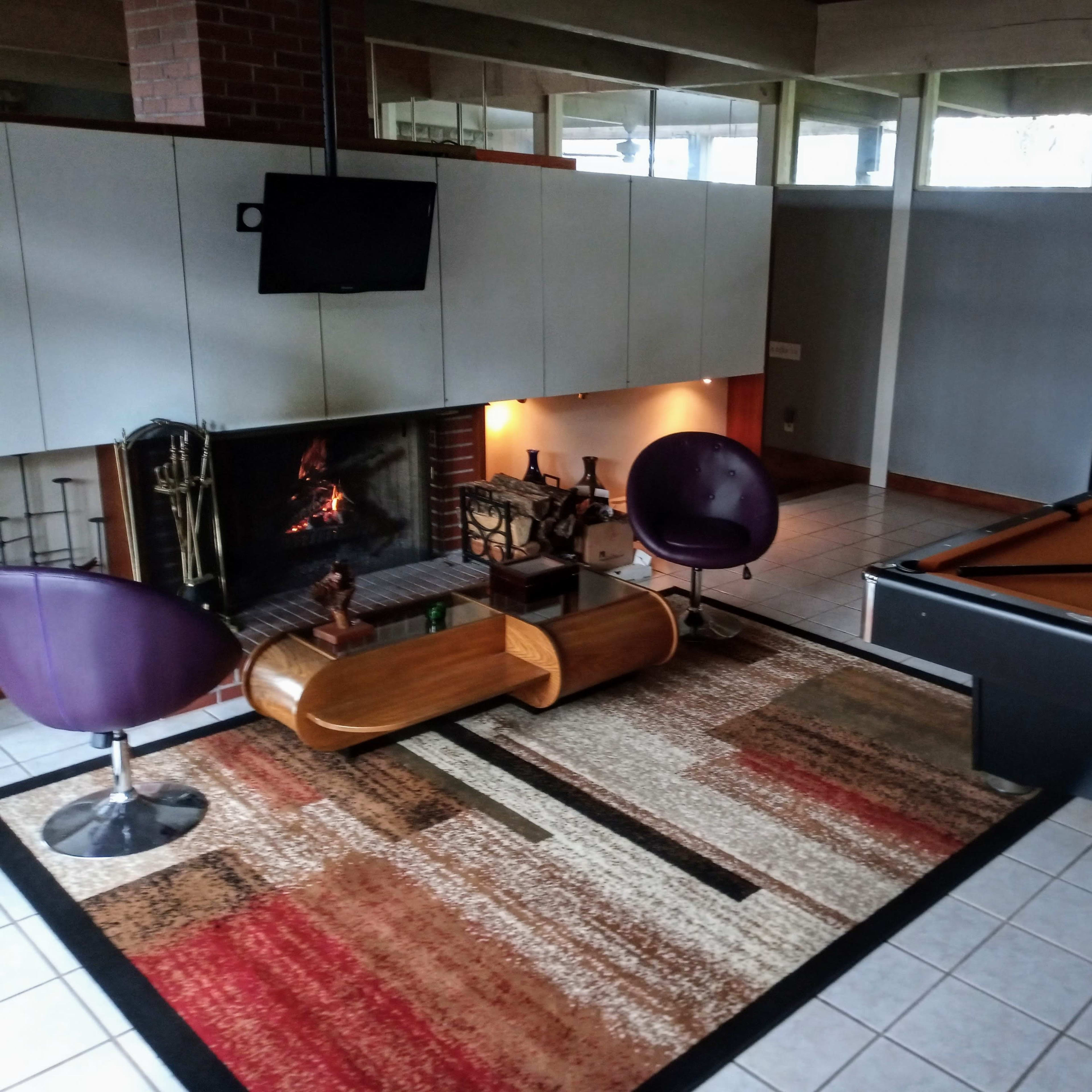 A game room featuring a pool table, a contemporary fireplace with a mounted TV, and modern purple chairs arranged on a patterned rug.