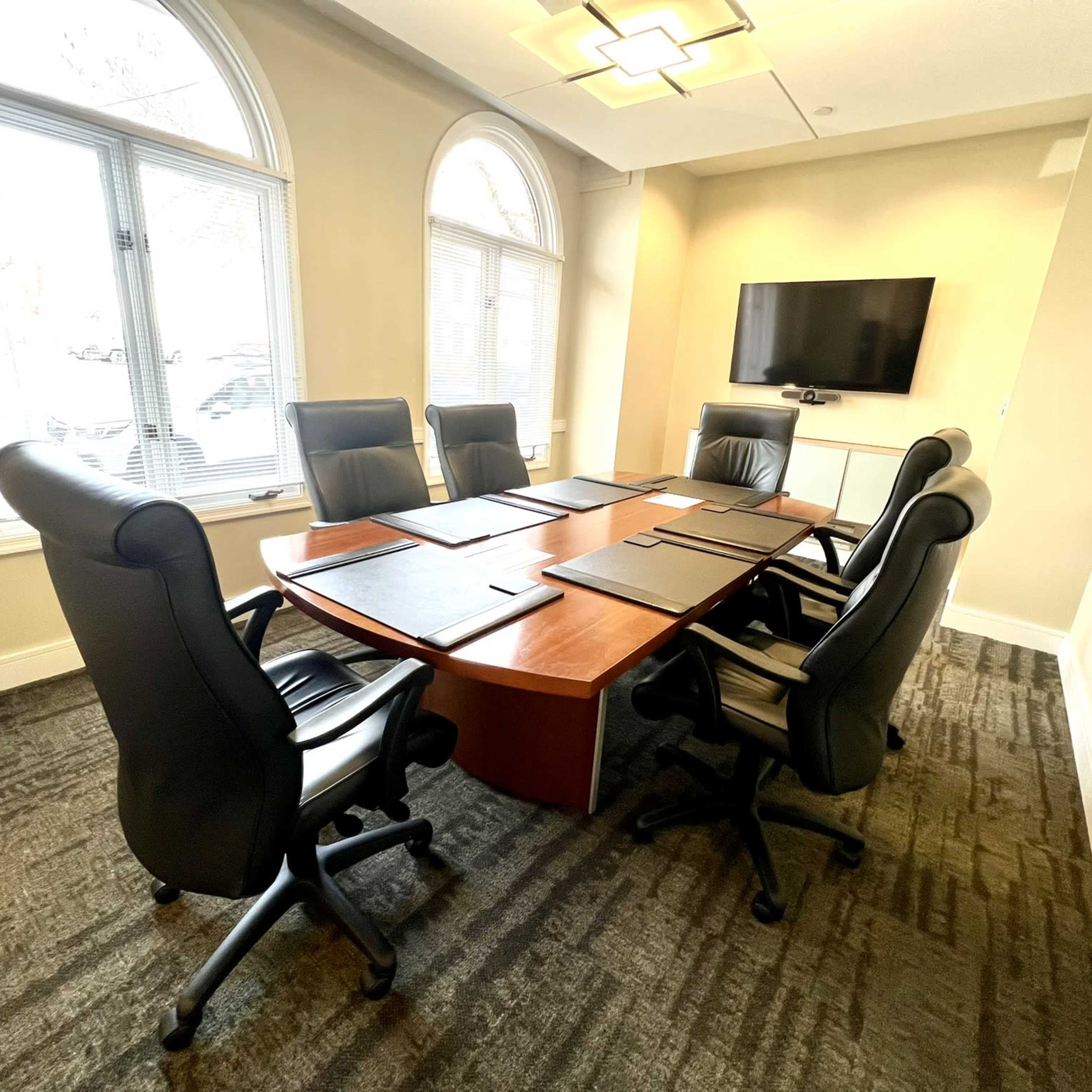 A conference room with a large wooden table surrounded by six black leather chairs and a wall-mounted television.