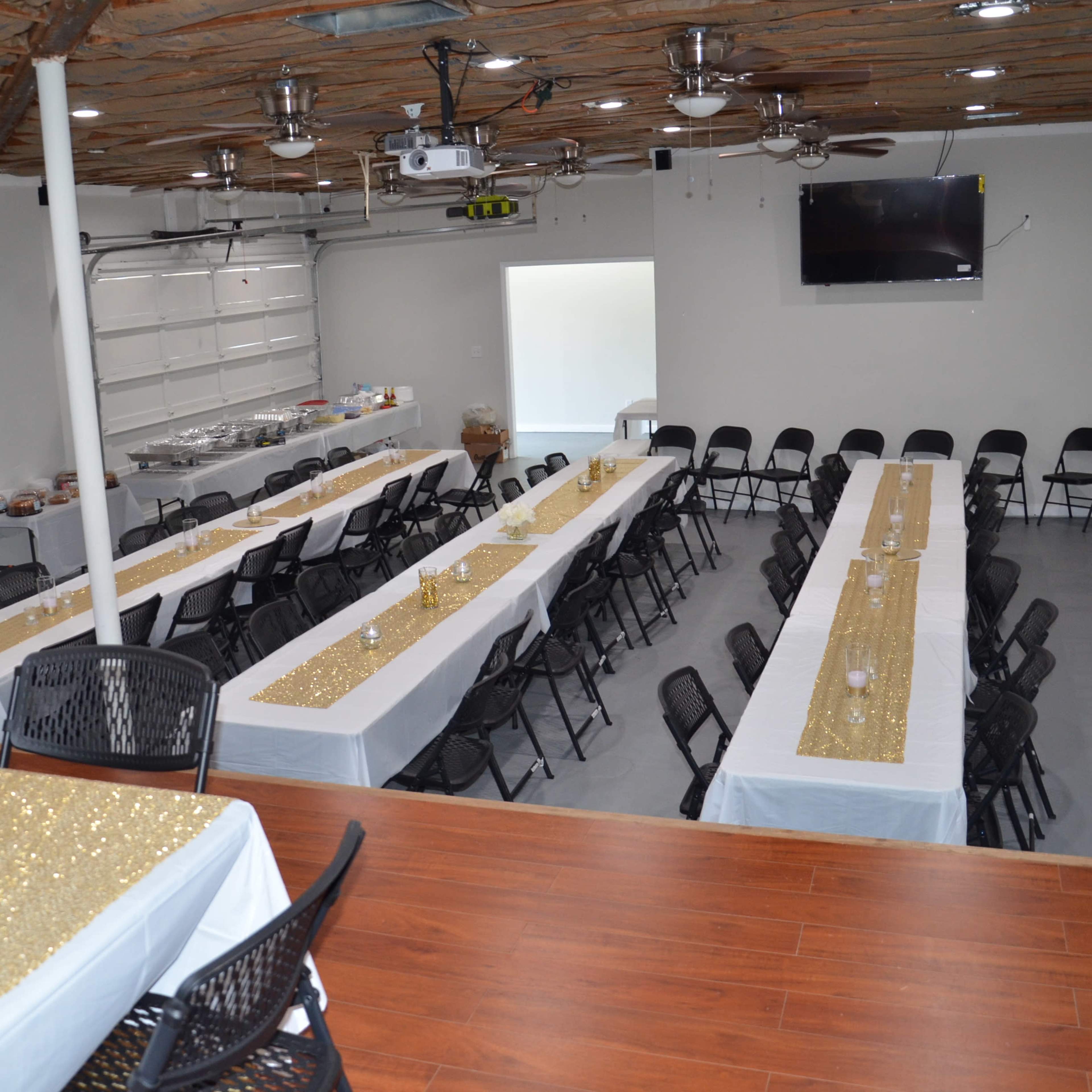 A spacious event room set with multiple long tables covered in white and gold tablecloths, surrounded by black chairs.