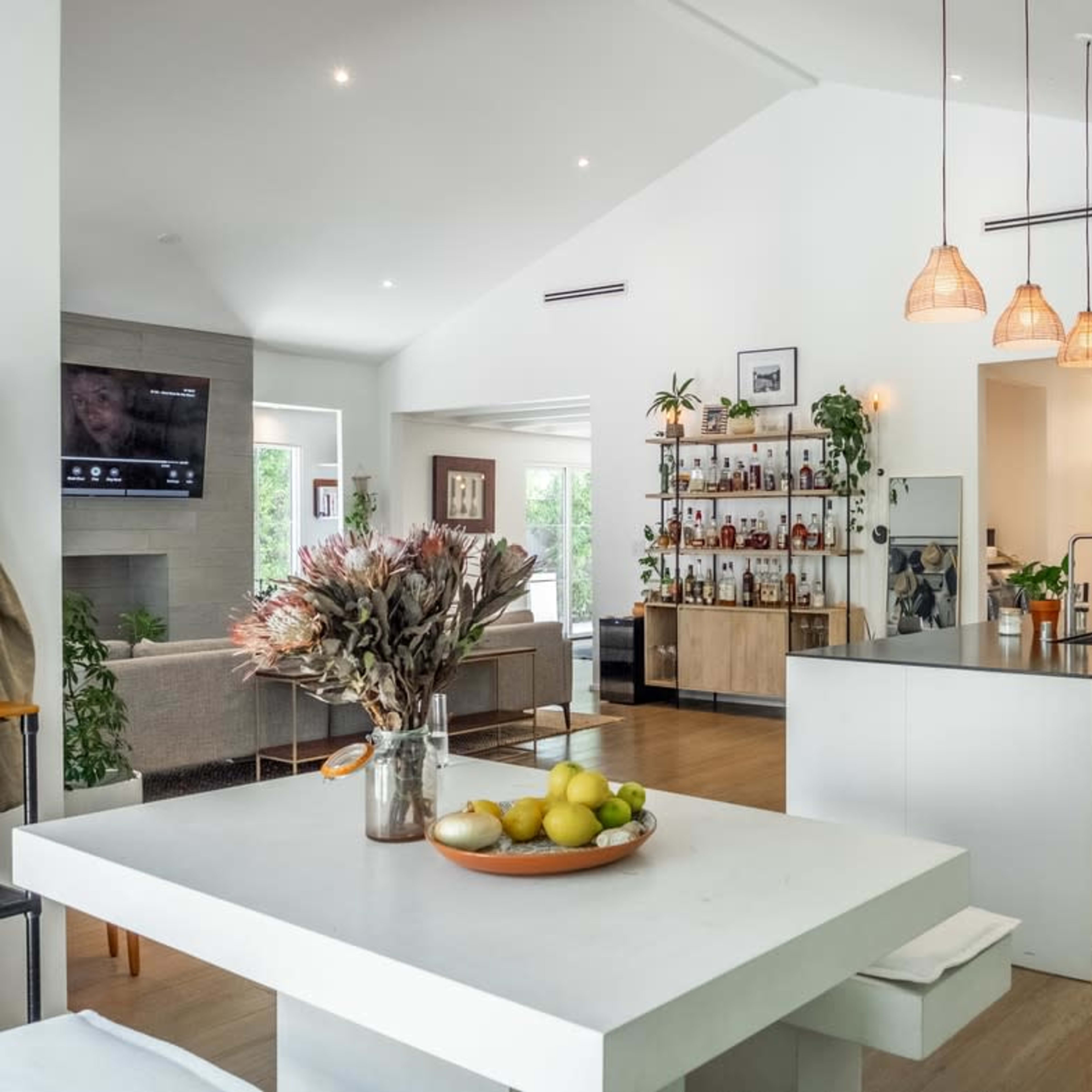 A modern kitchen features an island with a sink, hanging pendant lights, and open shelving filled with bottles and plants, adjacent to a seating area.
