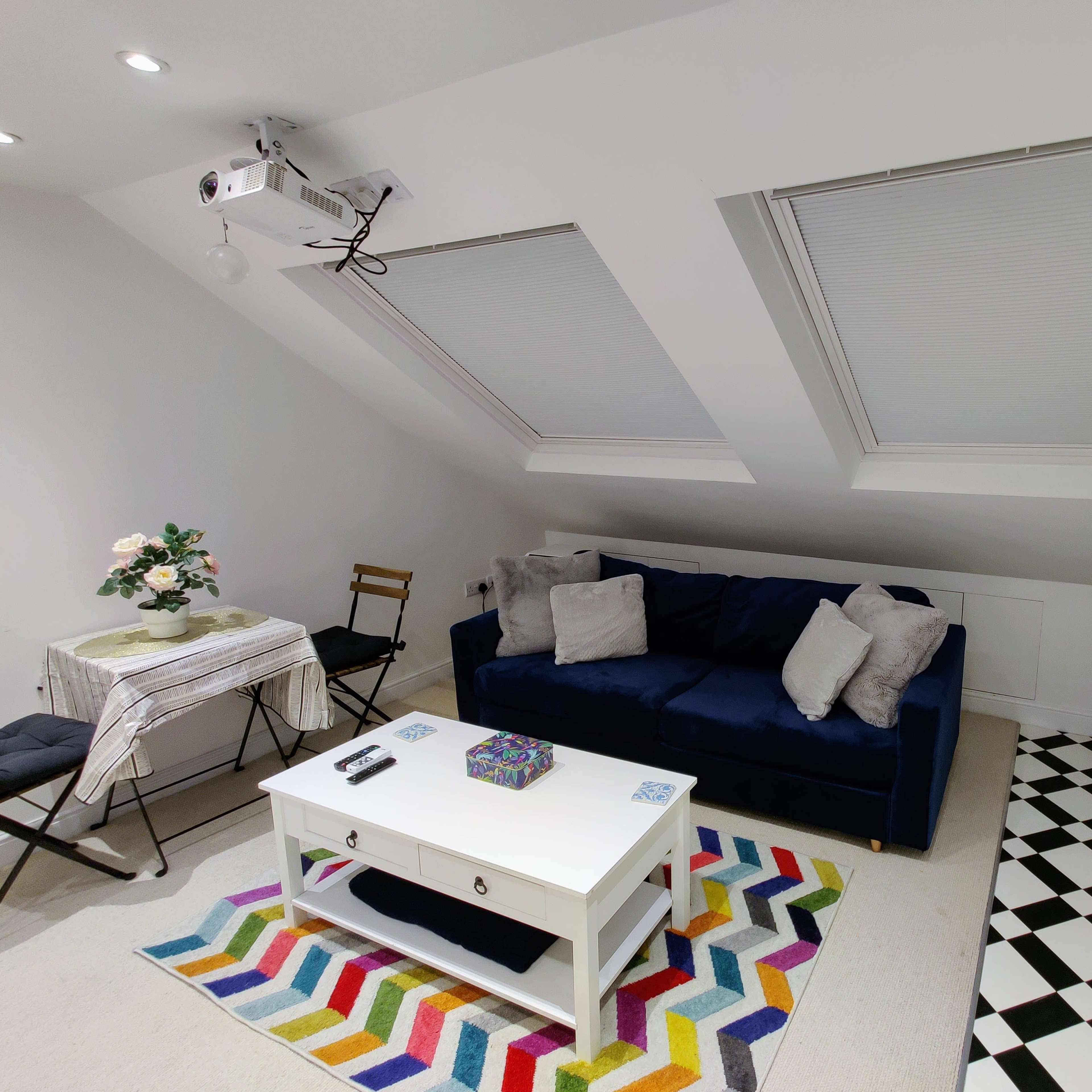The room features a blue sofa, a white coffee table, a colorful rug, and large skylight windows, with a kitchenette and dining area visible.
