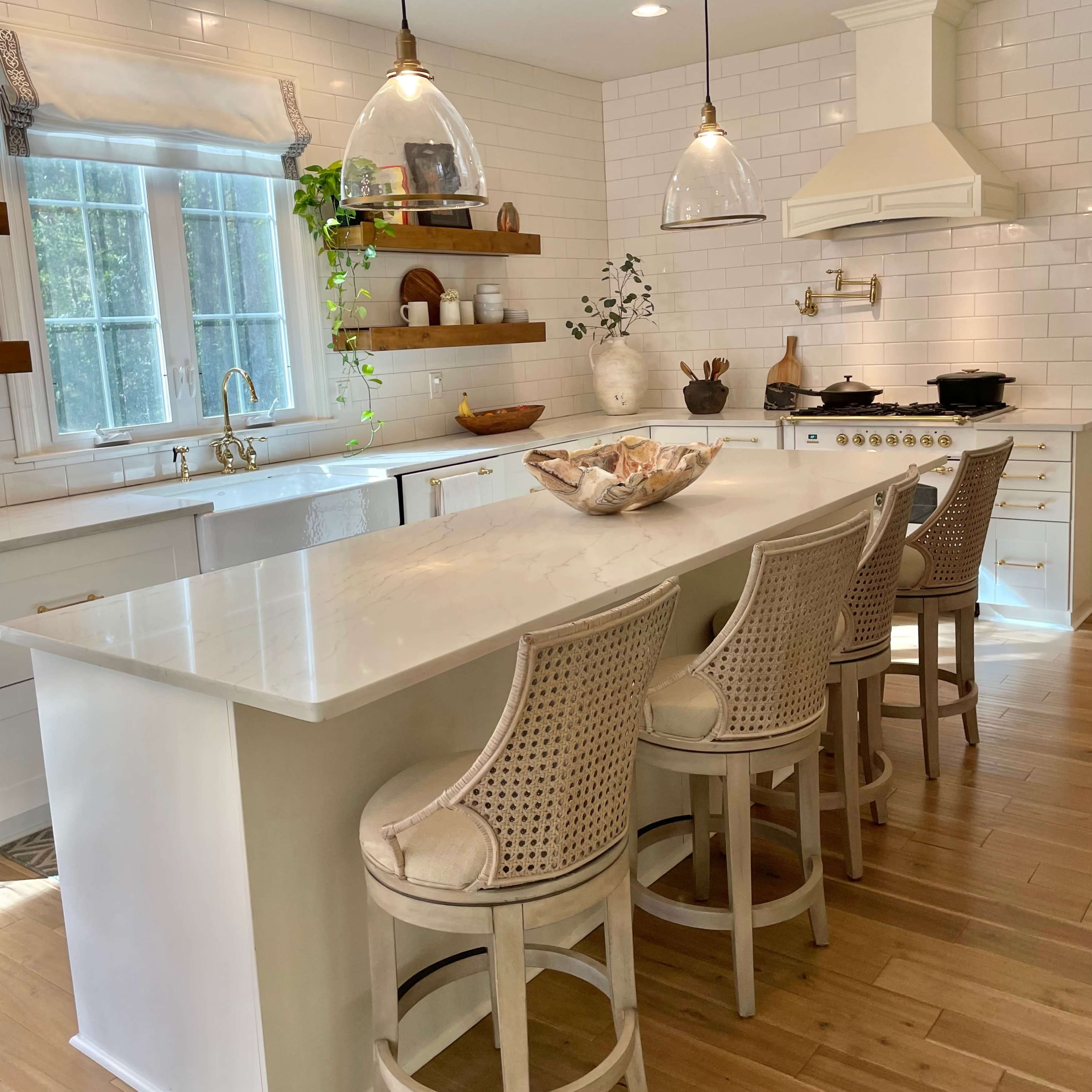 A modern kitchen features a large white island with four rattan bar stools and a window with natural light, accented by pendant lights and a stove against tiled walls.