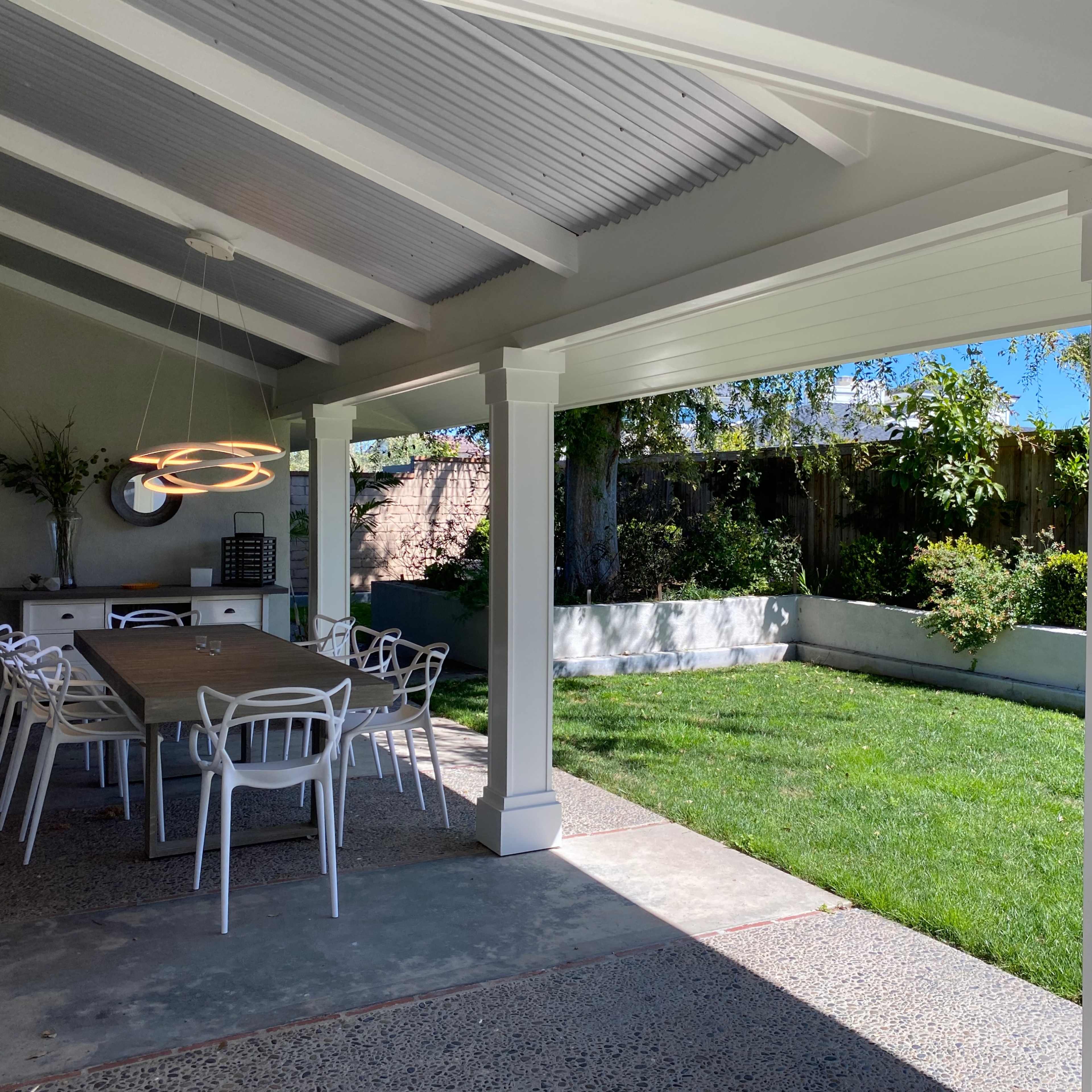 A covered outdoor dining area features a long table surrounded by white chairs, with a grassy lawn and garden visible in the background.