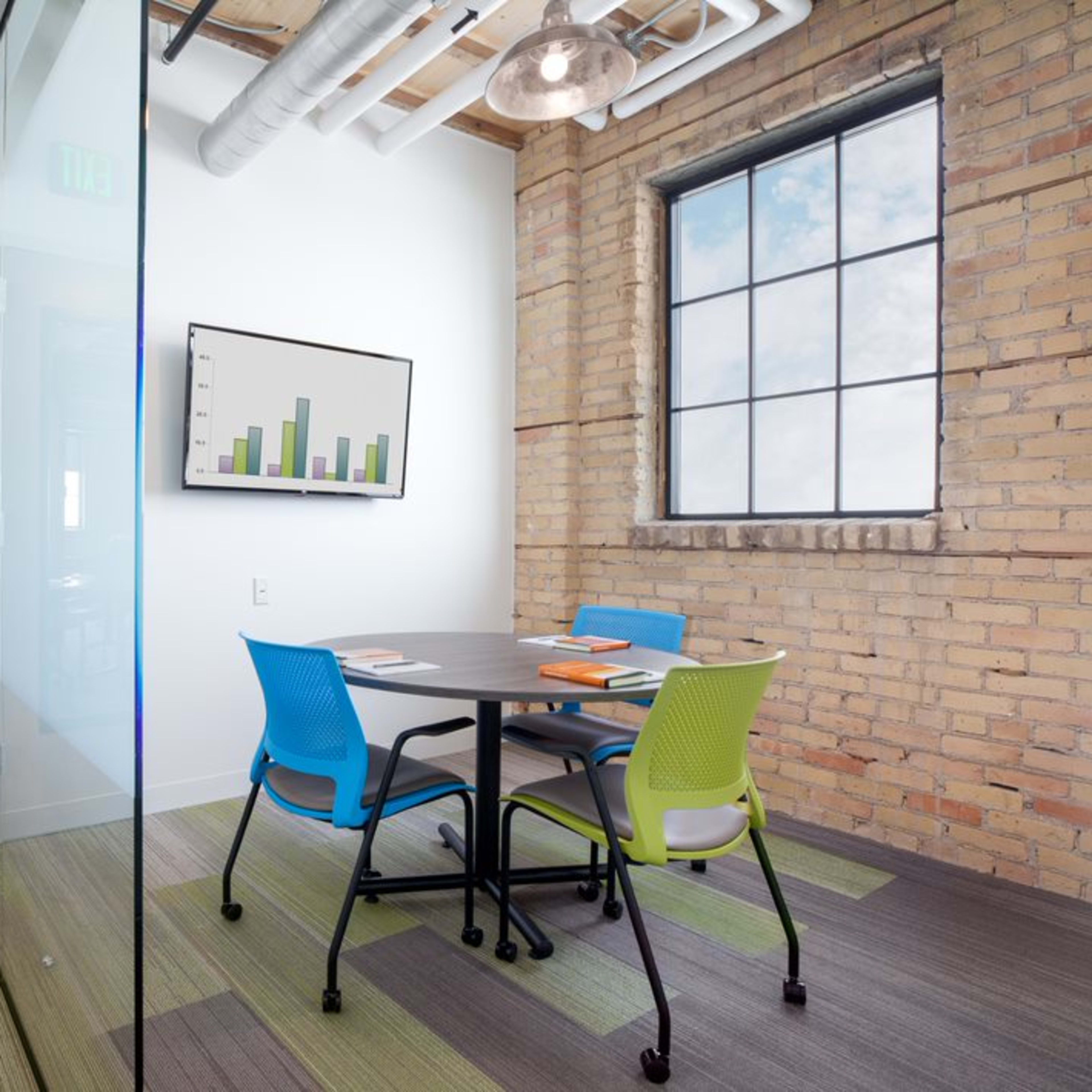 A small meeting room with a round table and colorful chairs, situated near a glass wall and a door marked with an exit sign.