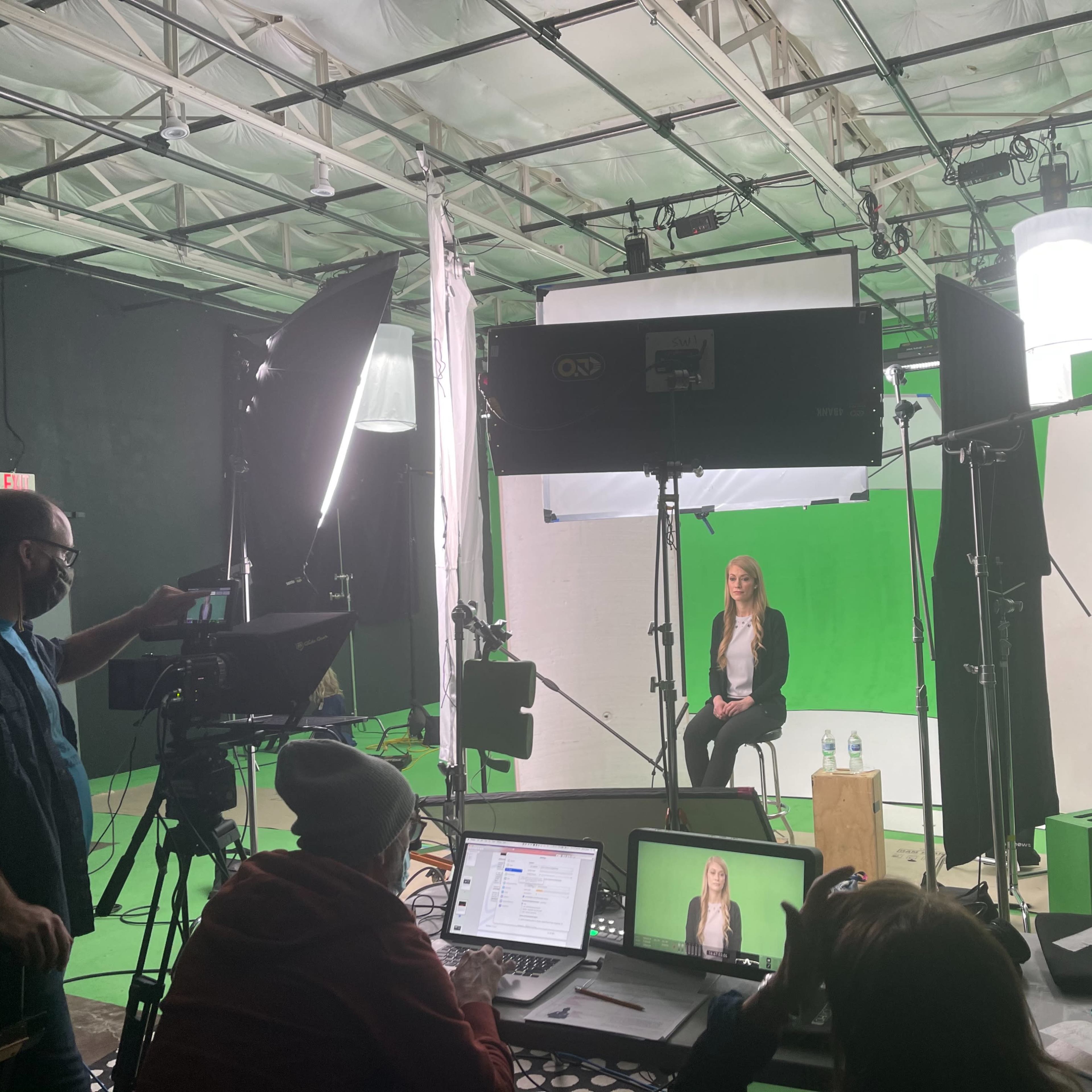 A film crew sets up equipment around a woman seated in front of a green screen in a studio.