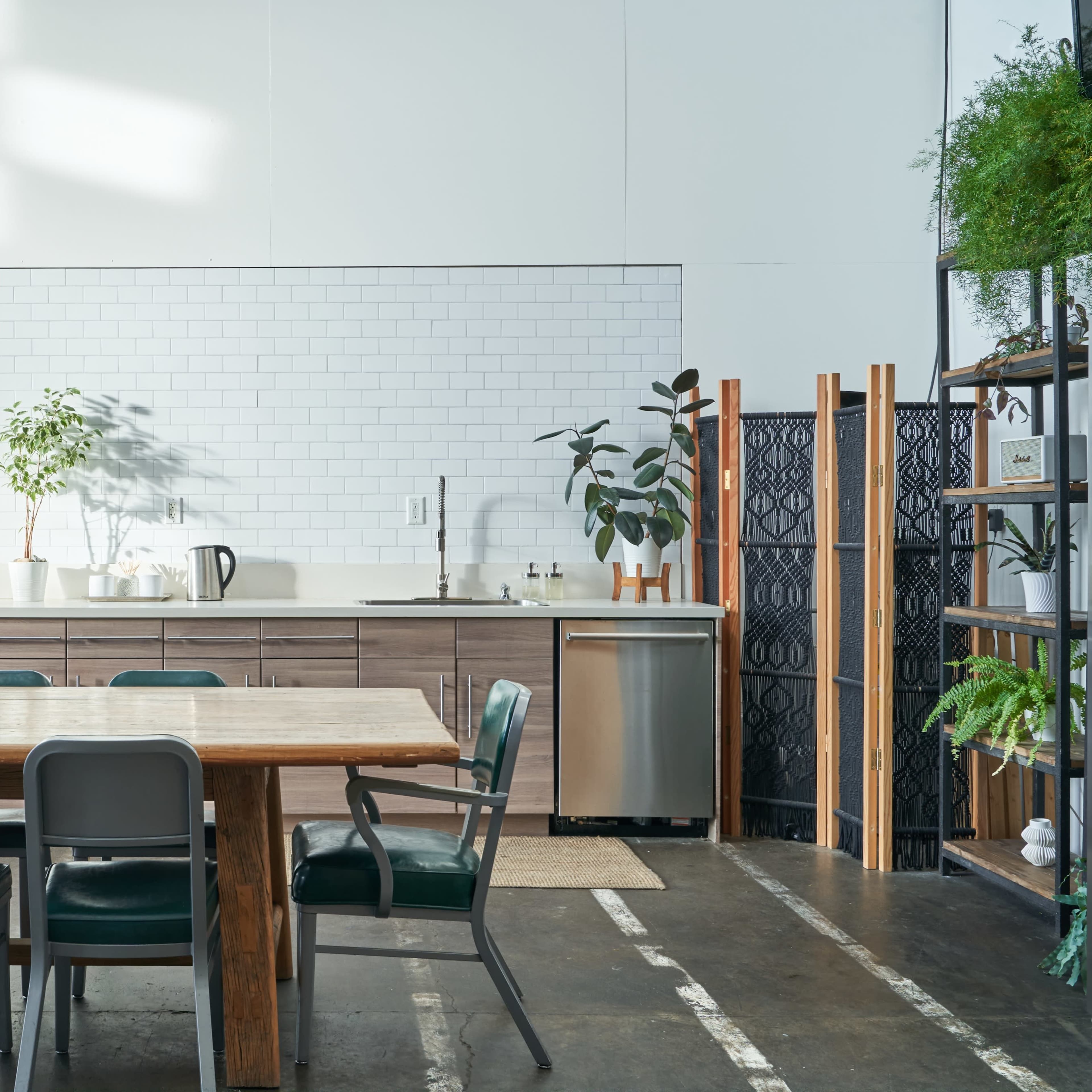 A modern kitchen space with a wooden dining table, metal chairs, potted plants, and a stainless steel appliance setup against a white tiled wall.
