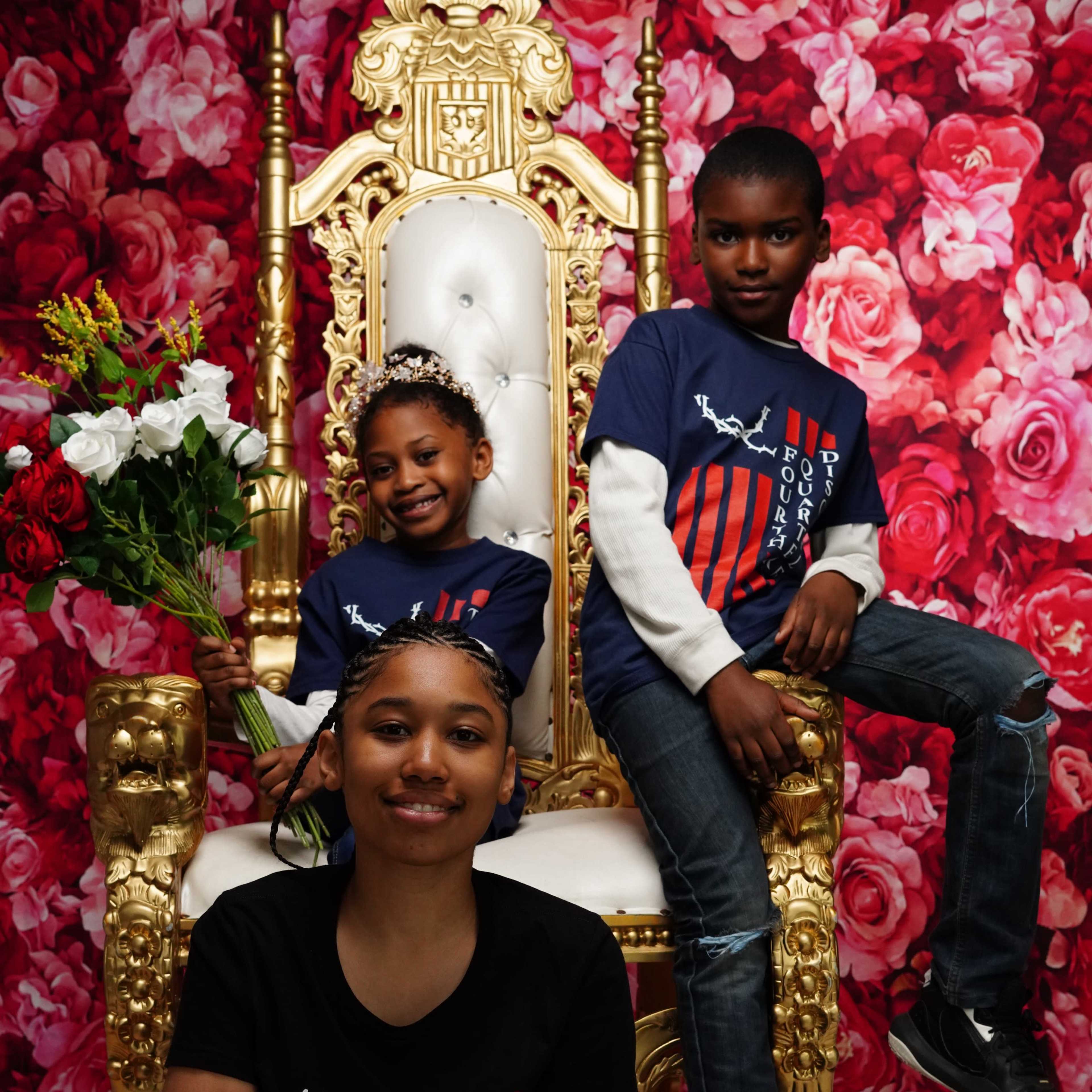 Three children pose in front of a floral backdrop, with one sitting on the floor and two others standing and seated on a golden throne, holding flowers.